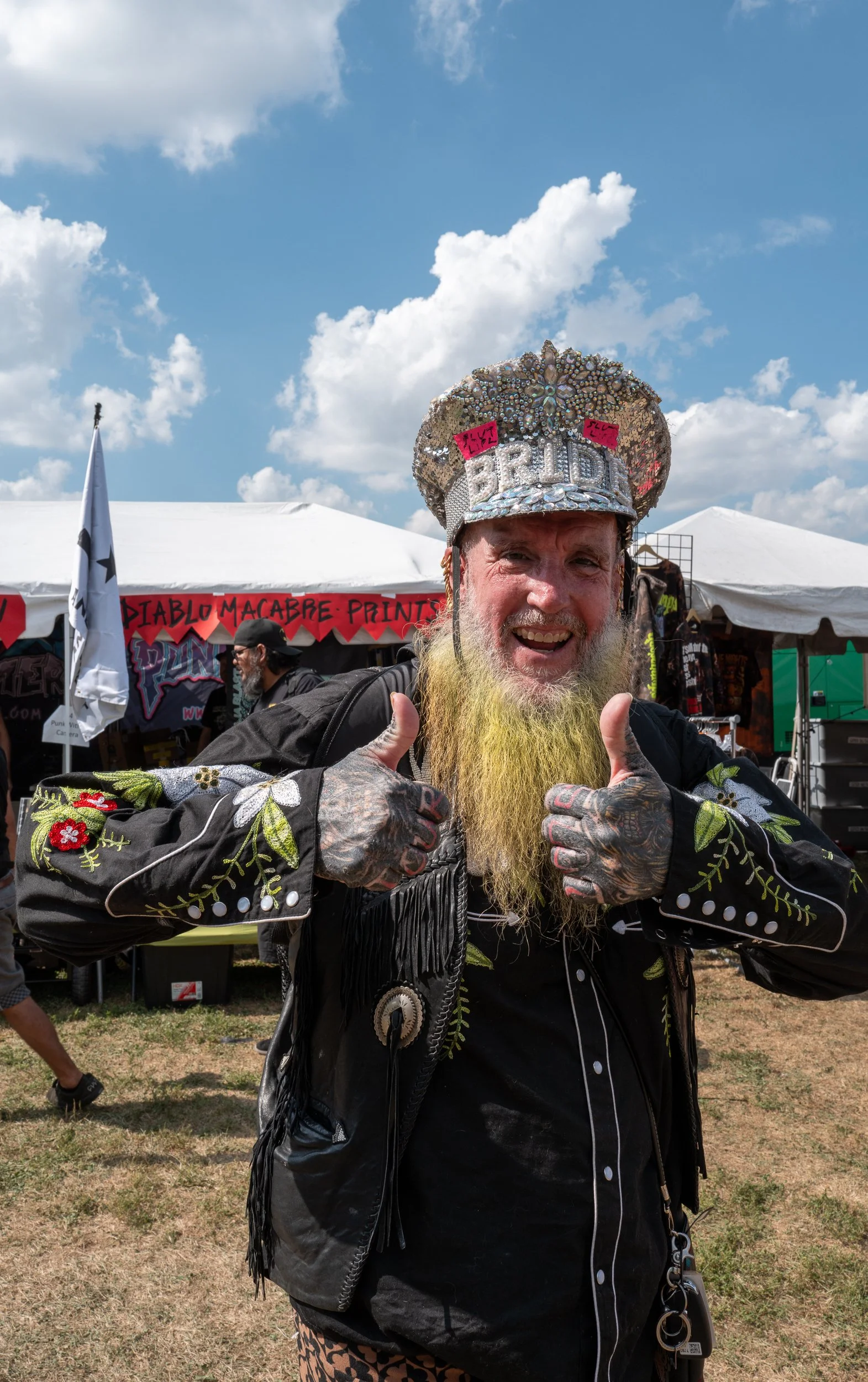 A man with a long yellow beard wearing a shiny decorated hat and giving two thumbs up at an outdoor event with tents and banners in the background.
