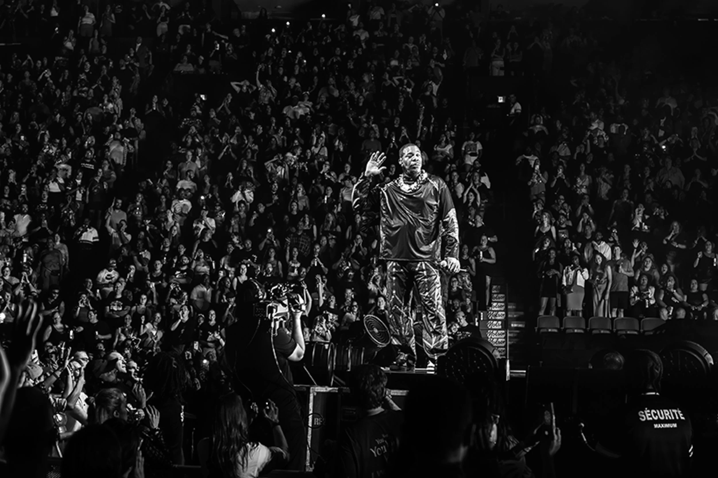 A person standing on stage with a microphone, raising their hand, in front of a large crowd in an indoor arena.