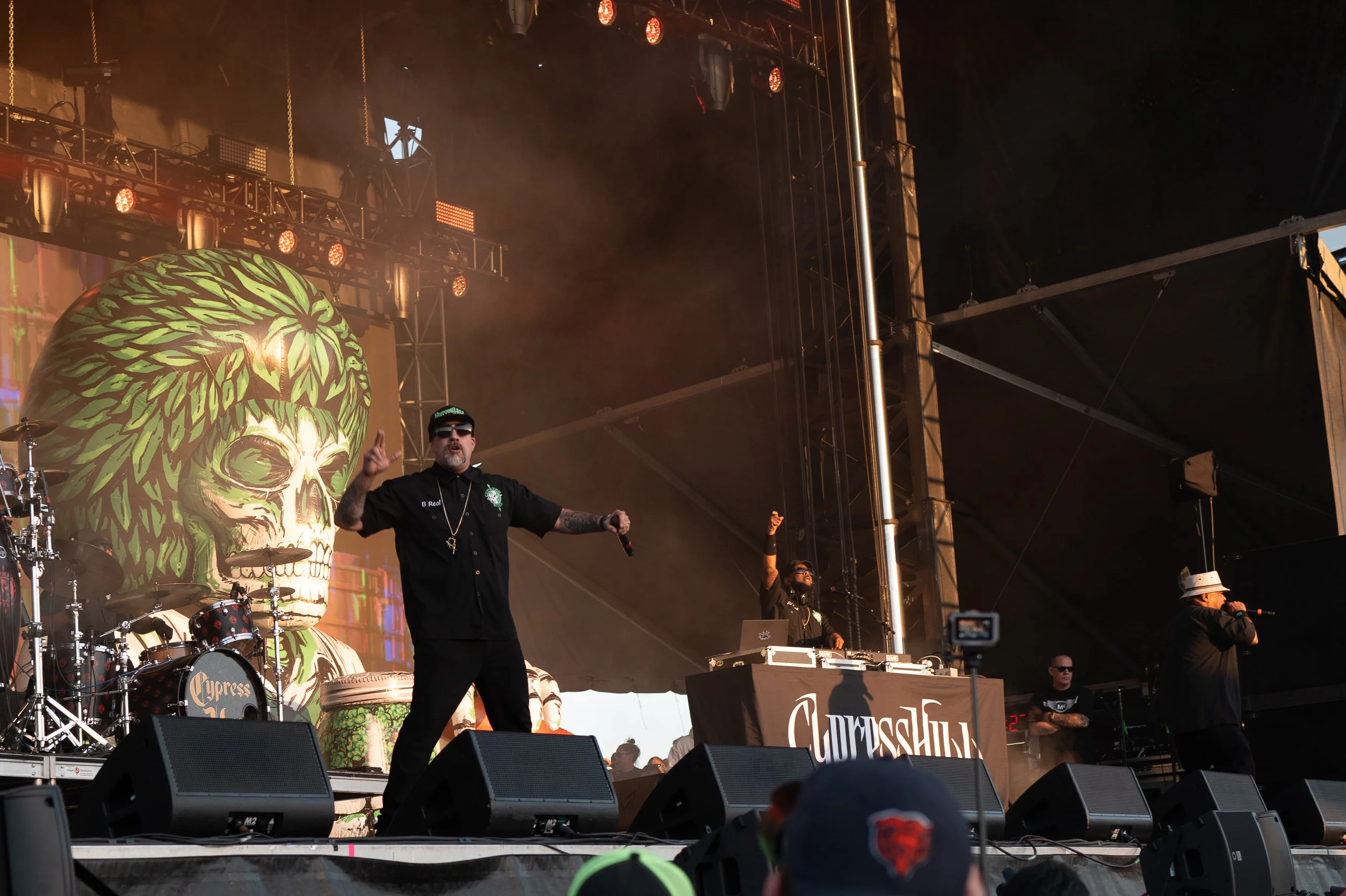 A live music performance on a stage with a DJ and two vocalists. The background features a large, artistic skull with green leaves and a watermelon pattern. The stage has various musical instruments and sound equipment.
