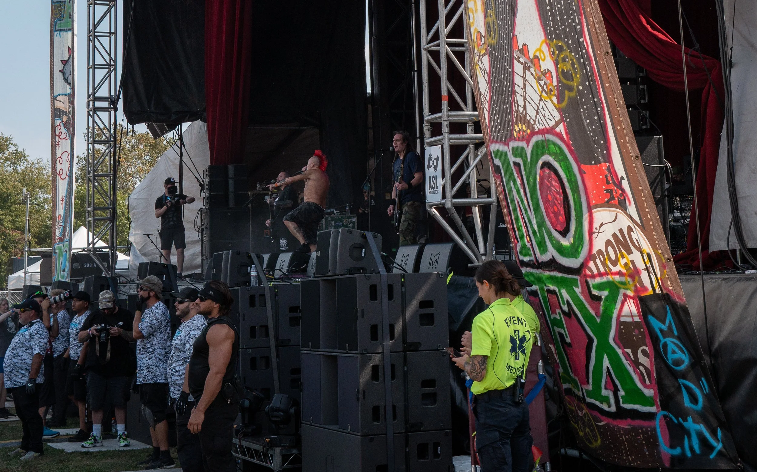 A live music performance on an outdoor stage, with a shirtless drummer with a red mohawk, a guitarist singing into a microphone, and stage crew members in the foreground. The scene includes a large colorful banner with graffiti-style art and the word