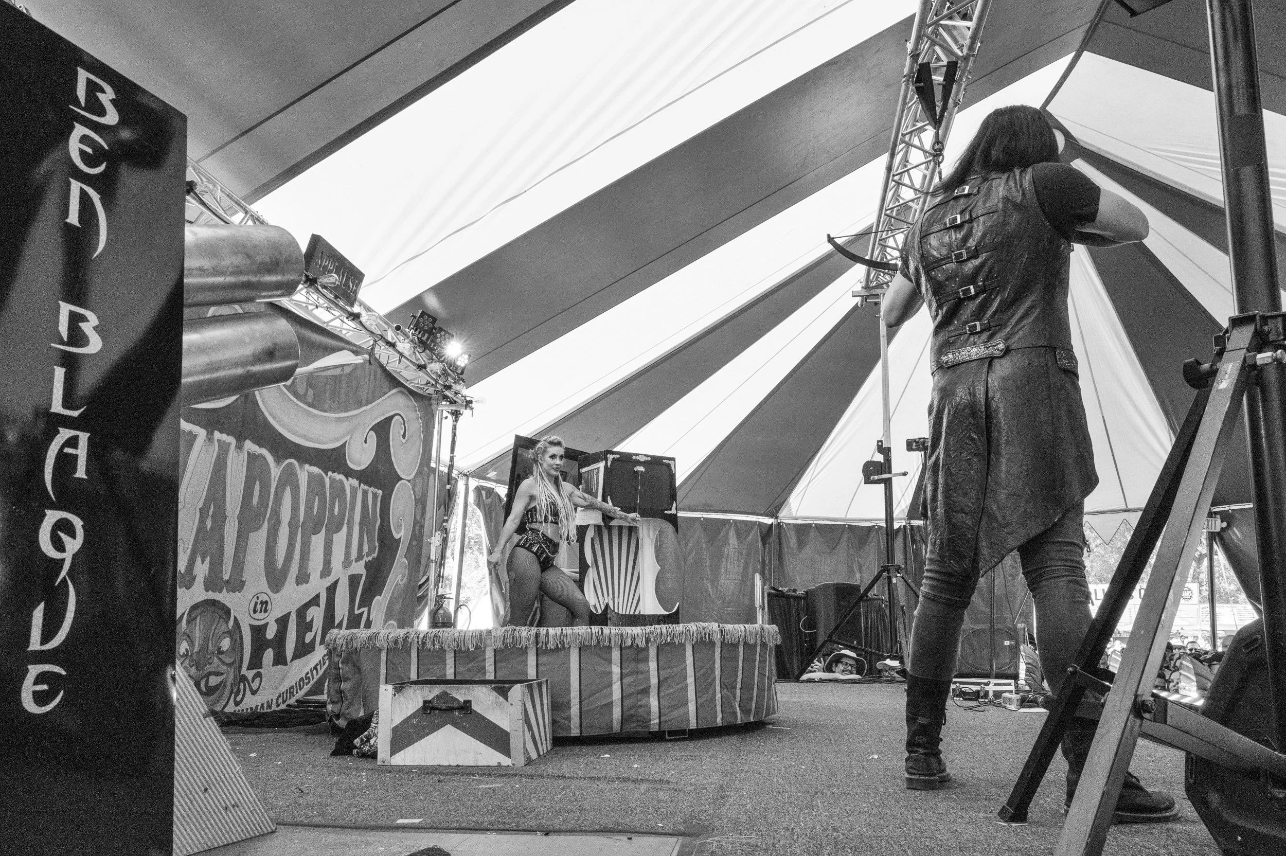 A performer on a small circus stage inside a tent, being photographed by a person in a leather vest. The stage has a colorful sign with the words "Circus in Hell" and other carnival-themed decorations.
