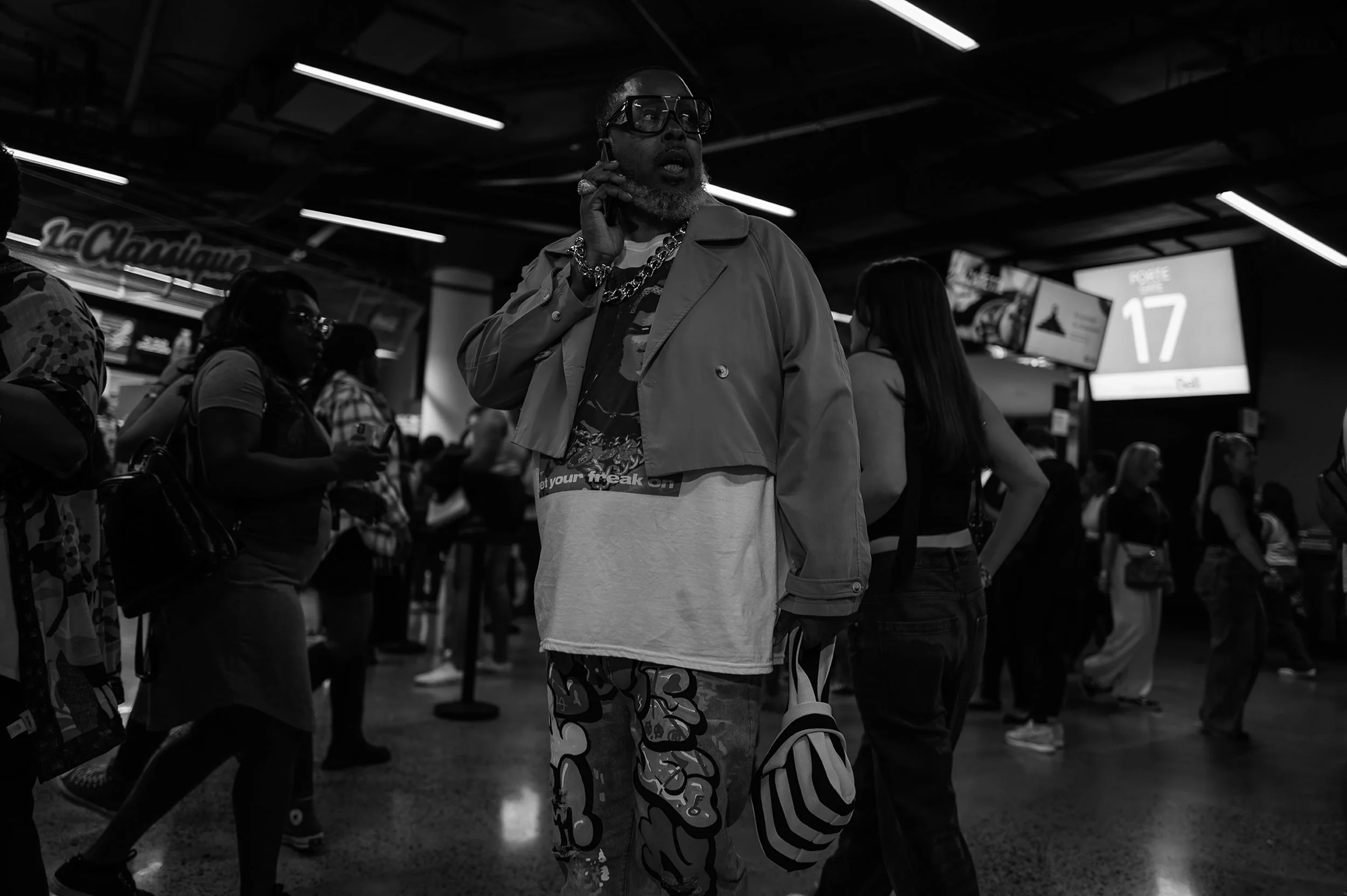 A man wearing glasses and jewelry holds a phone to his ear in an airport terminal, with other travelers visible in the background.