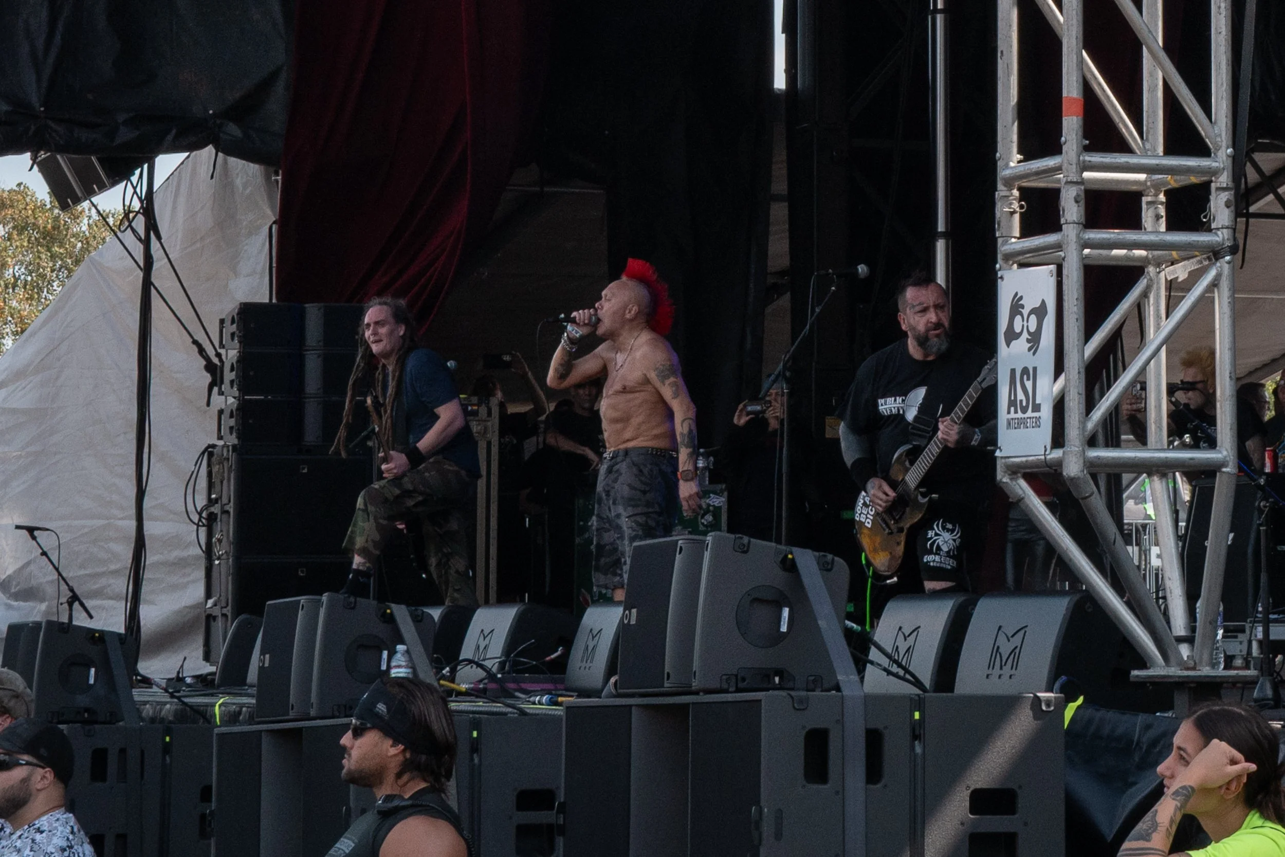 Band performing on stage at outdoor concert, singer with red mohawk shirtless, guitarist with black shirt, audience watching.