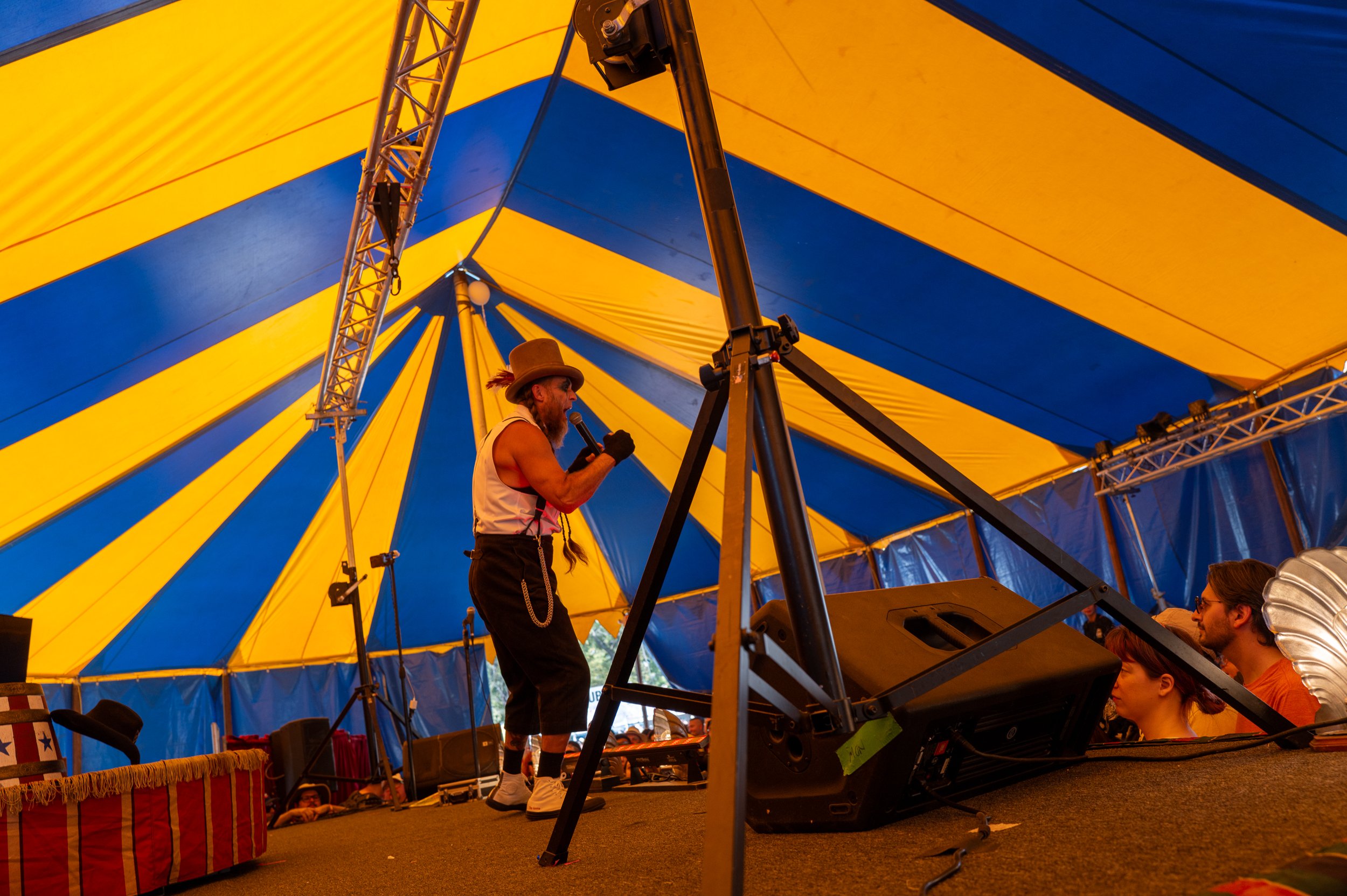 A performer with a beard, wearing a wide-brimmed hat, sleeveless shirt, and suspenders, singing into a microphone on a stage inside a yellow and blue striped tent during a circus or festival.