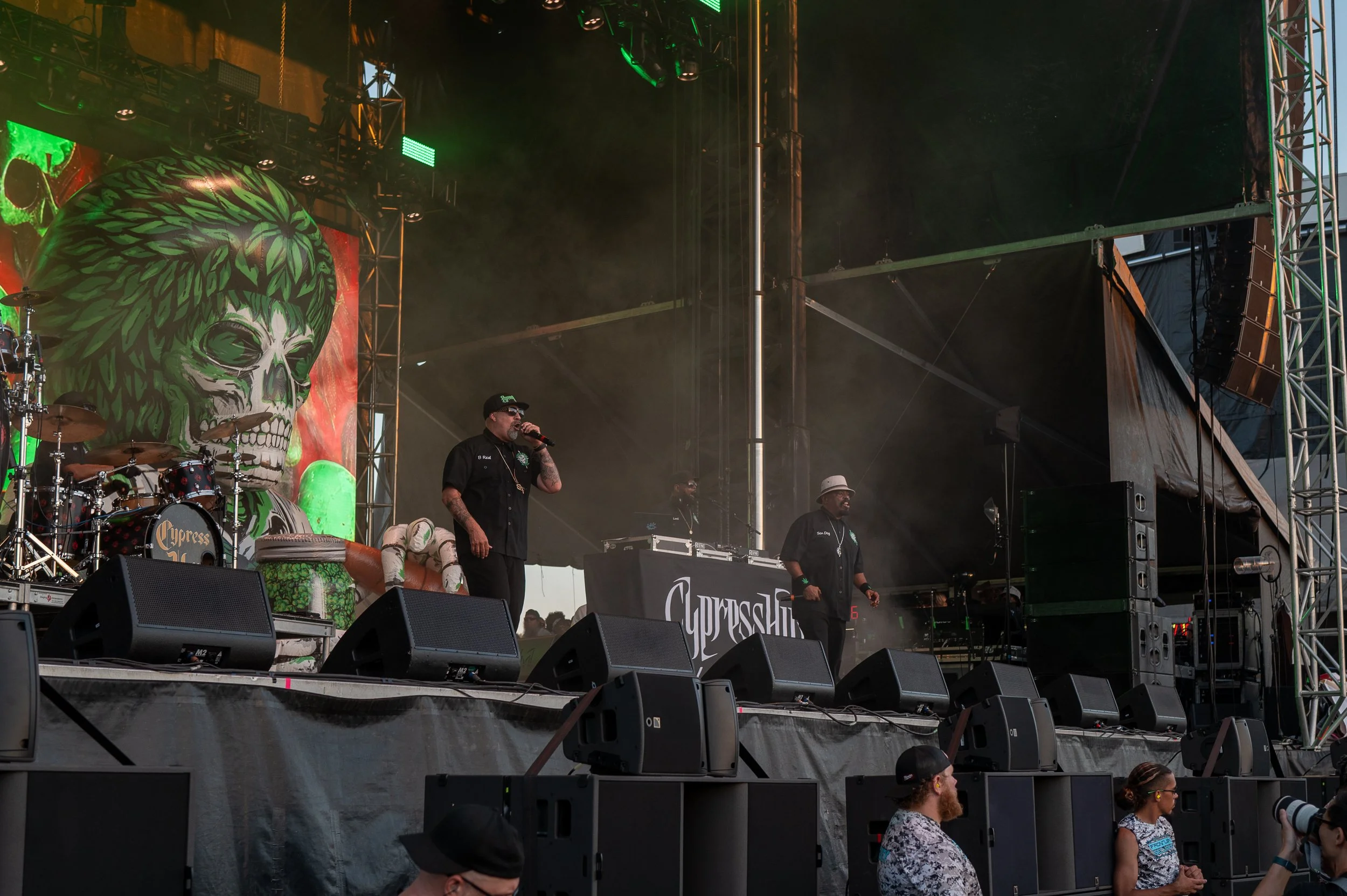 A band performing on stage at an outdoor event, with a large banner displaying a green and white skull with leaves in the background. Two men are singing or speaking into microphones, and there is a DJ setup behind them. The stage has multiple speake
