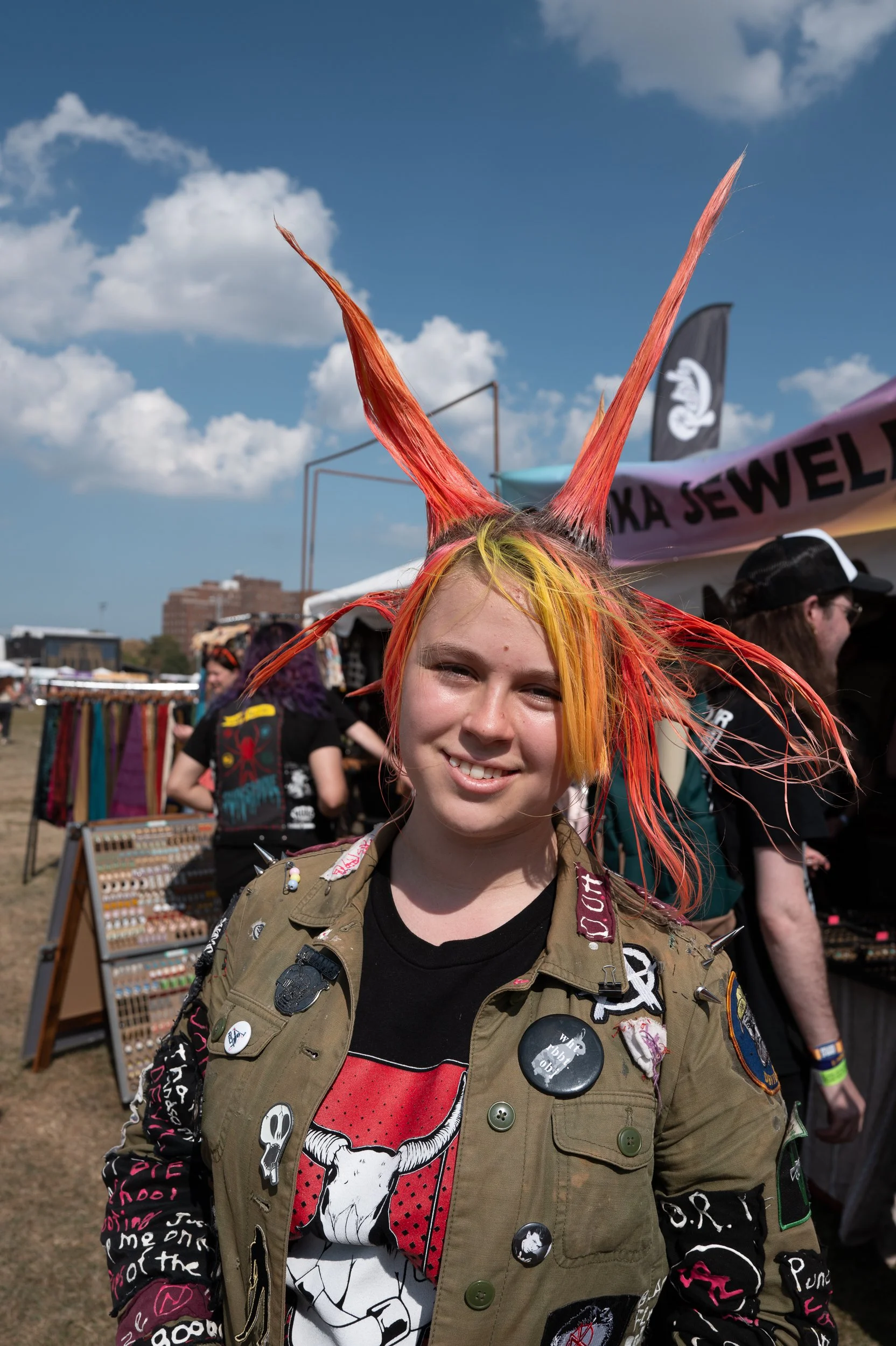 A young woman with bright orange and yellow hair styled into tall, spiked points, smiling at an outdoor festival or fair. She wears a military-style jacket adorned with various patches and pins, and stands in front of vendor booths and tents with a b