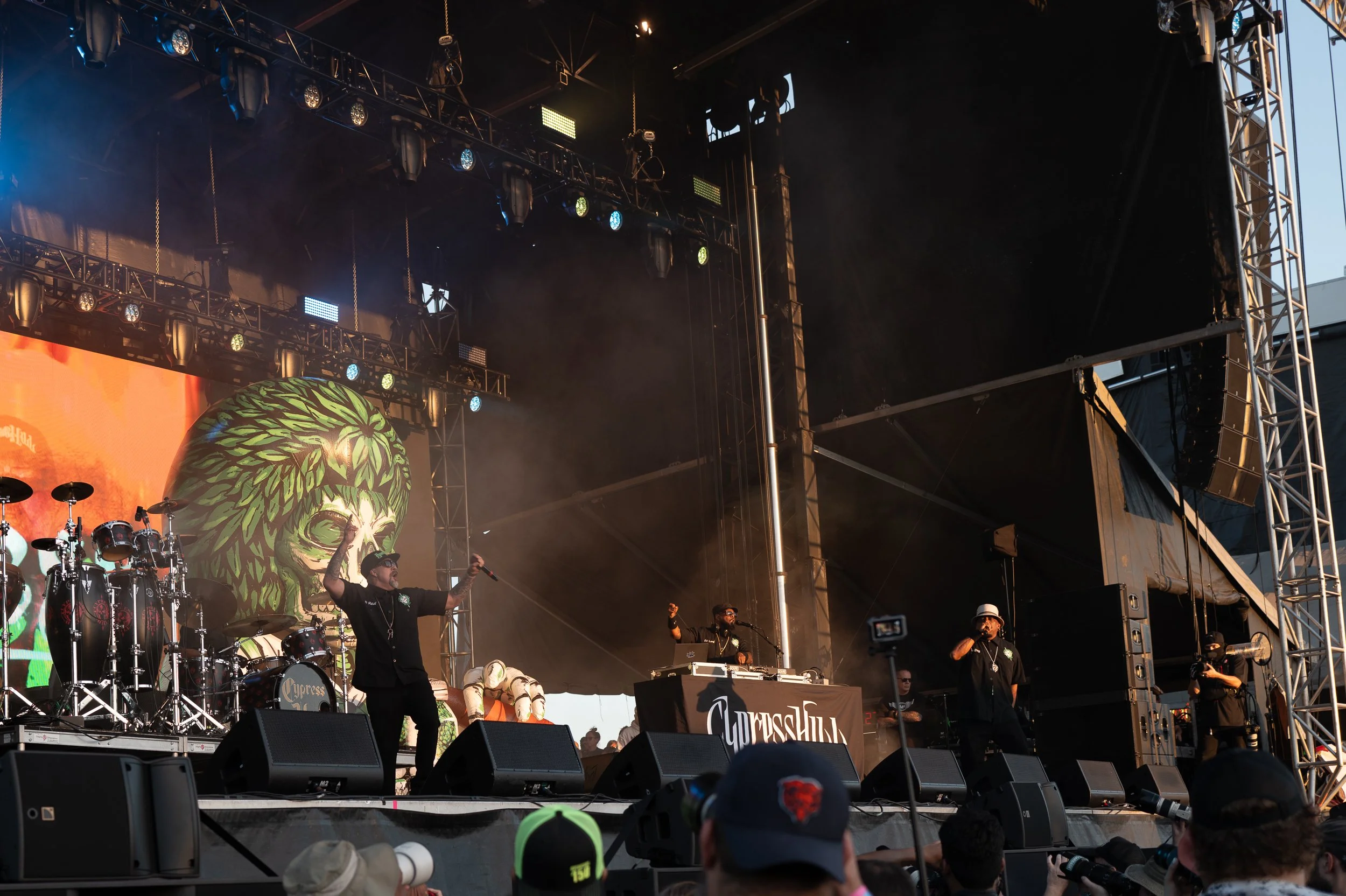 A live musical performance on an outdoor stage with a DJ and rappers, featuring a large LED screen with a green skull and foliage design in the background, audience members in the foreground, and sound and lighting equipment.