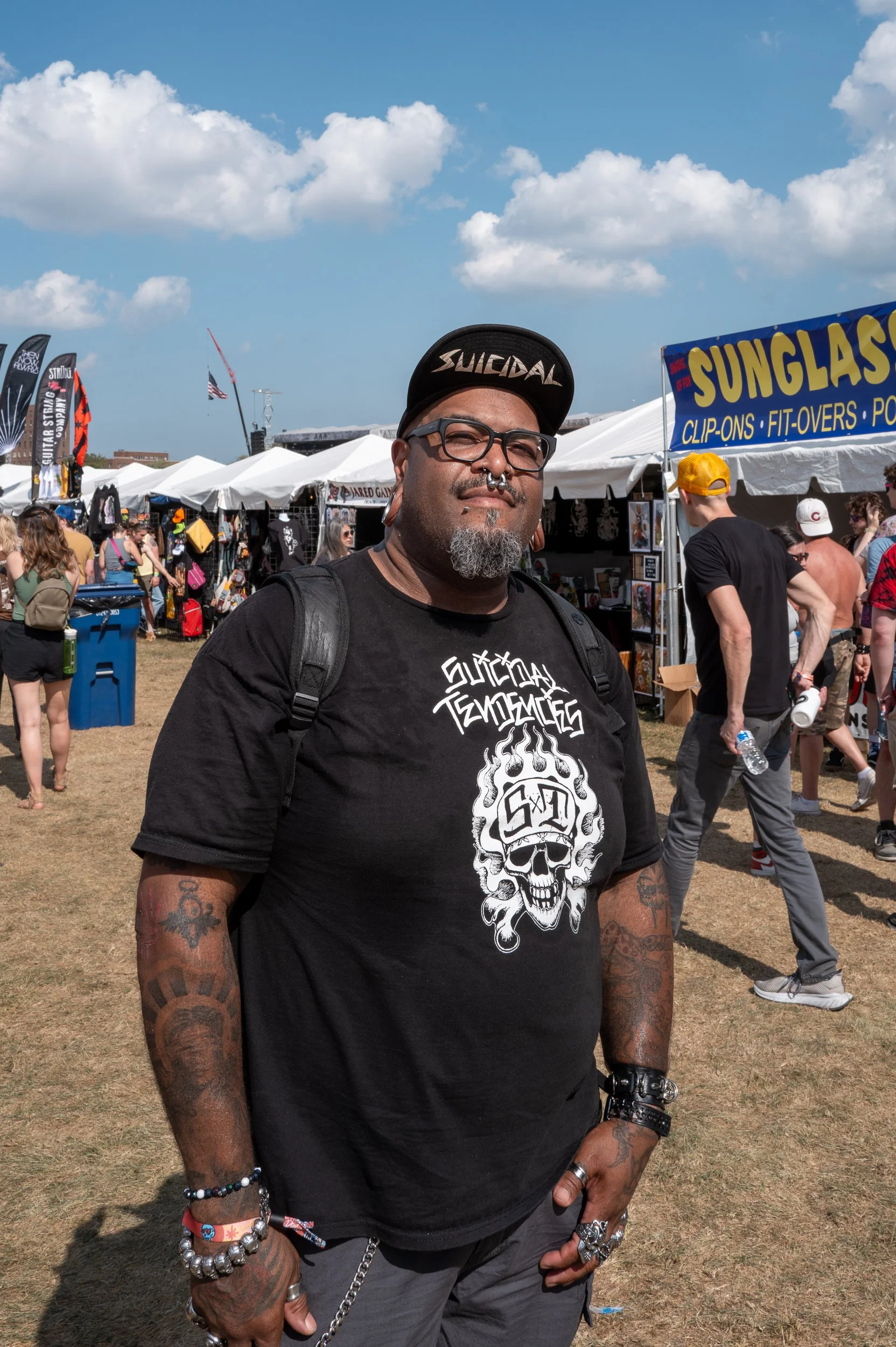 A man with tattoos, glasses, and a septum piercing stands outdoors at a festival with tents and people in the background, wearing a black graphic T-shirt and a black cap.