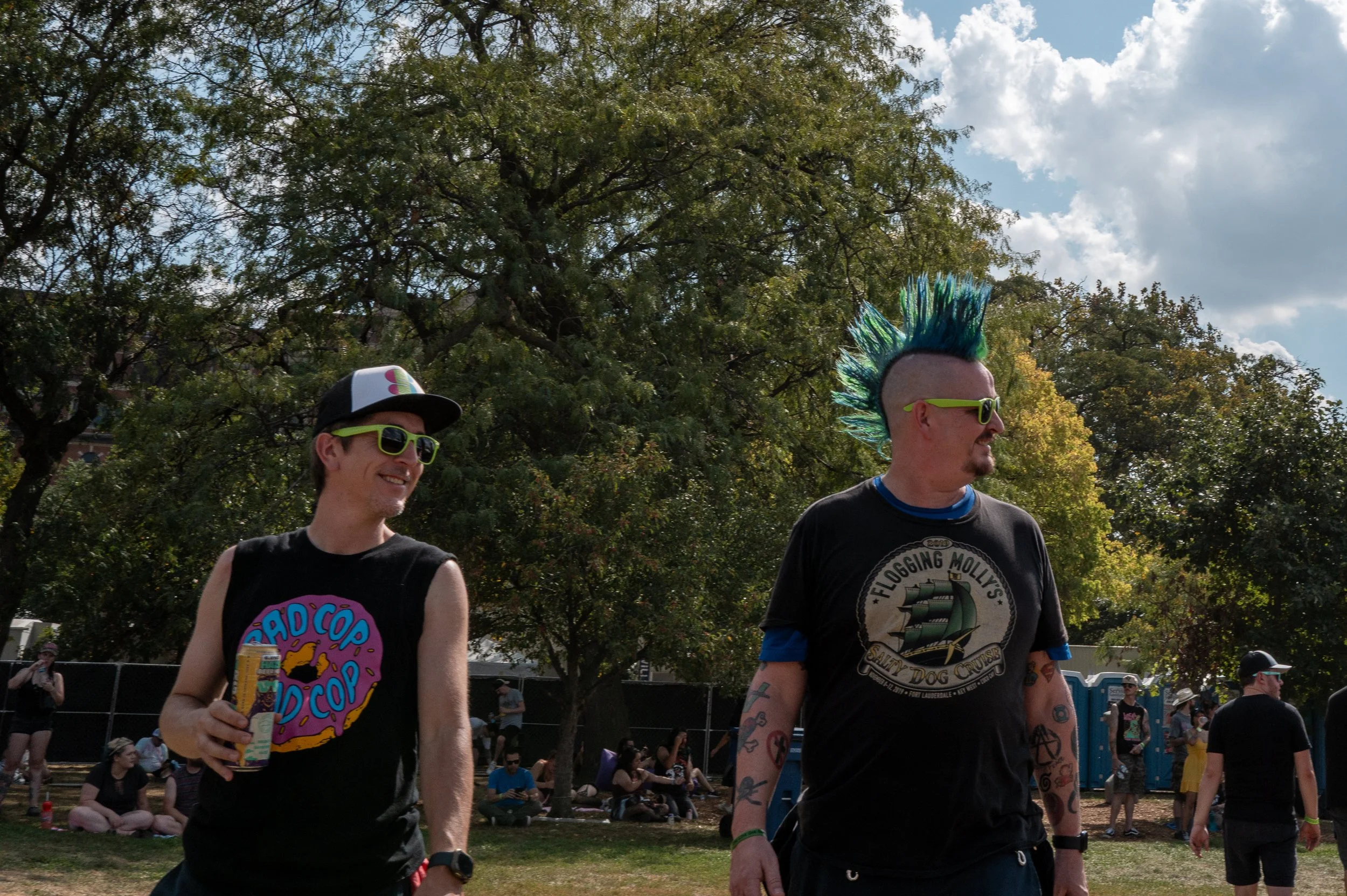 Two men with brightly colored hair and sunglasses enjoying an outdoor event with people sitting and standing around in a park.
