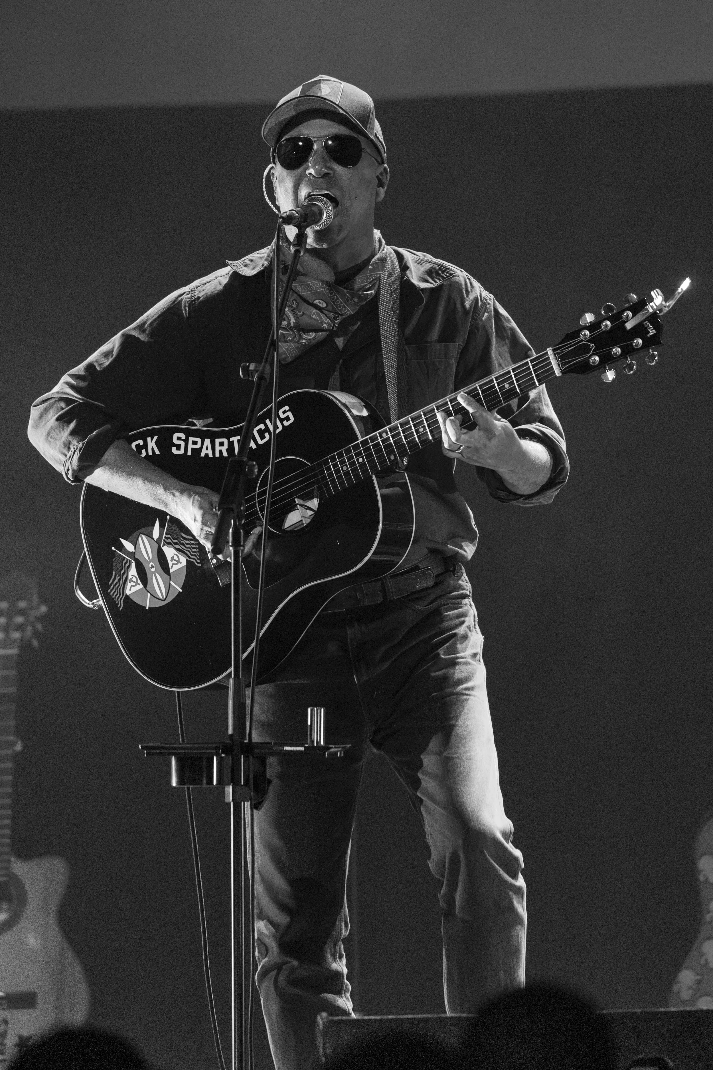 A man wearing sunglasses, a cap, and a bandana around his neck, singing into a microphone while playing an acoustic guitar on stage, black and white photo.