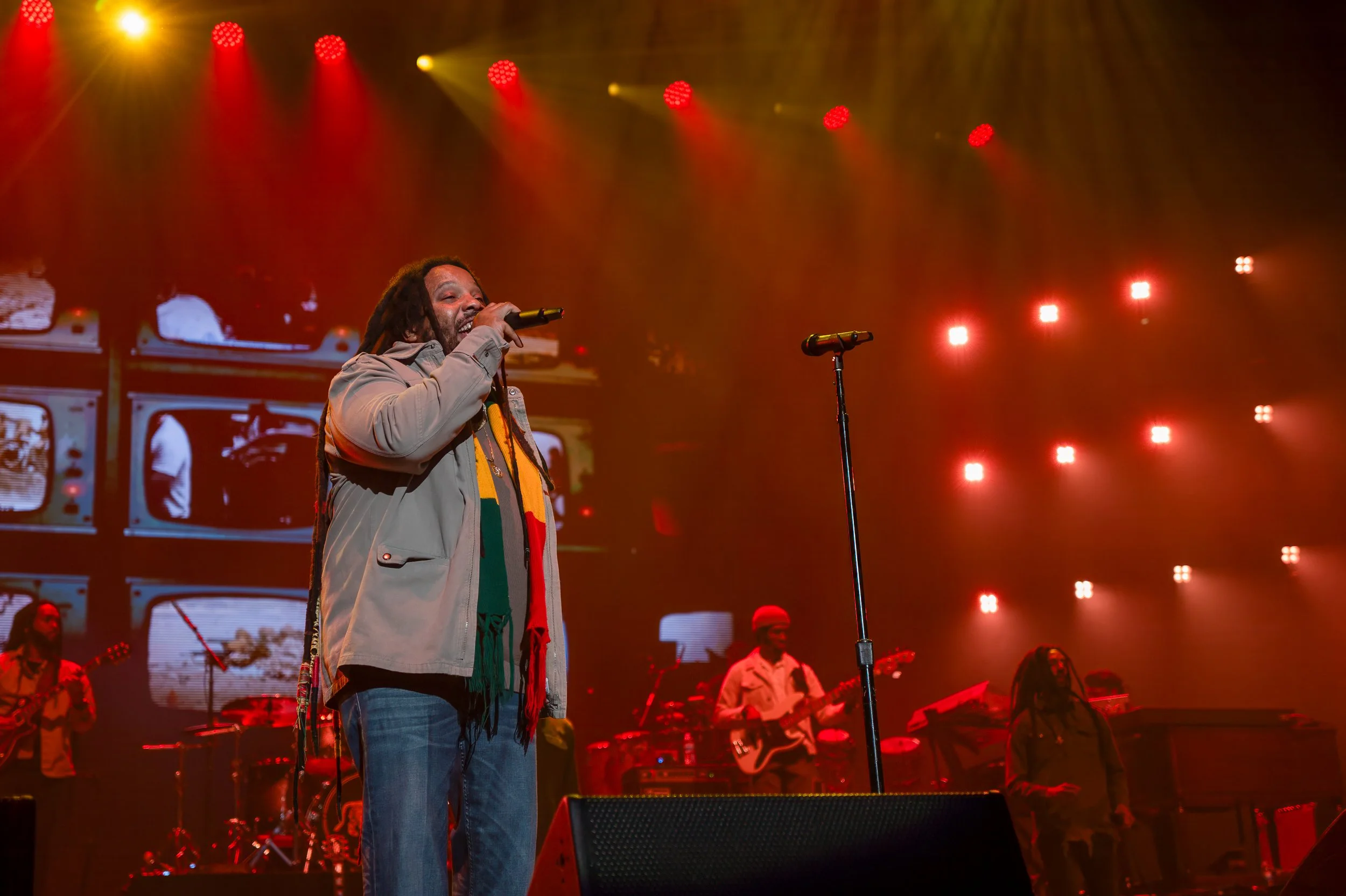 A male singer with dreadlocks performing on stage with a microphone, with a band playing instruments behind him, under colorful stage lights.