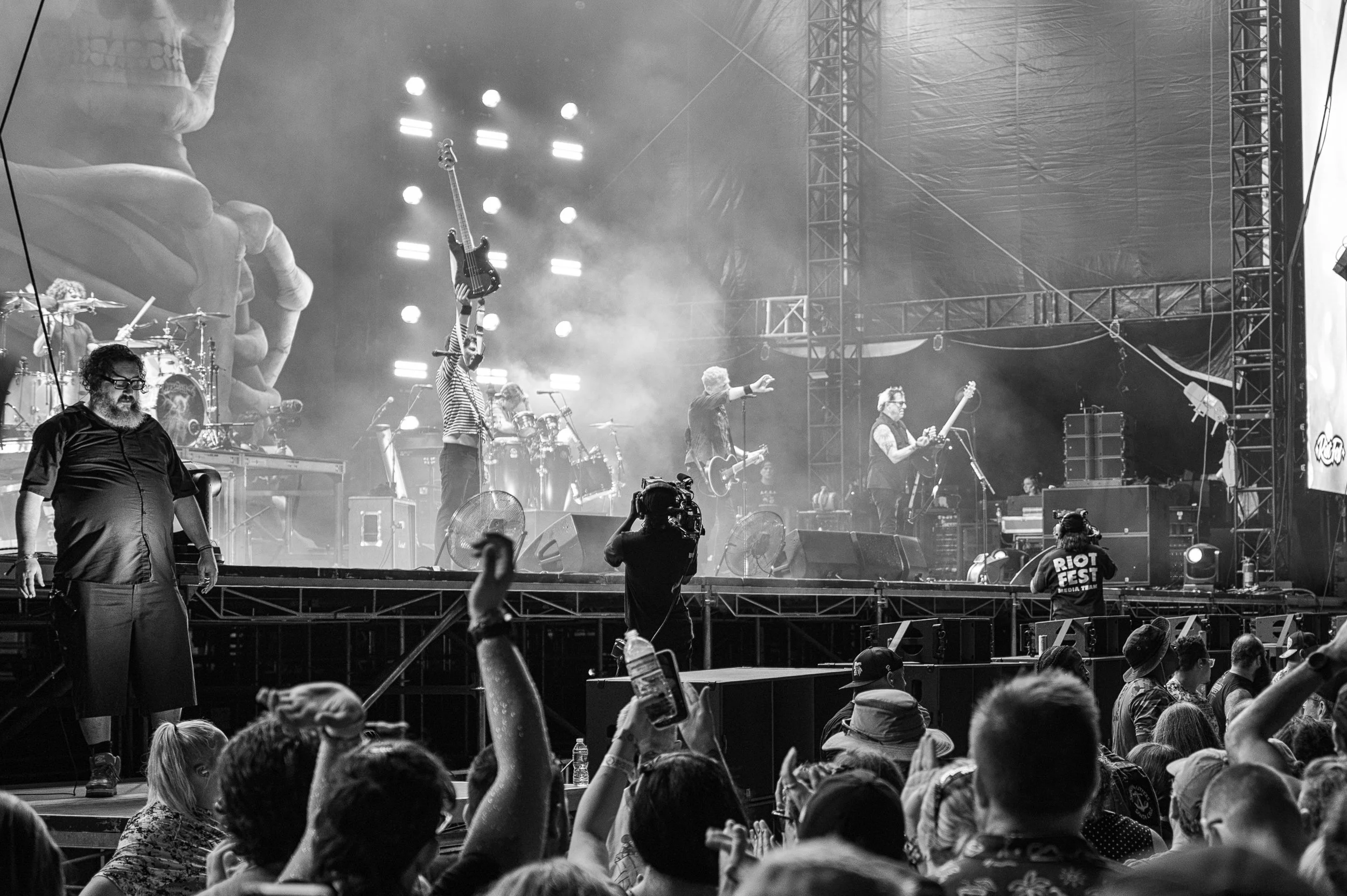 Black and white photo of a live music performance at a festival with a large crowd. The band is on stage with various instruments, and one member is holding a guitar upright. Audience members are raising their hands and holding drinks, and there is a