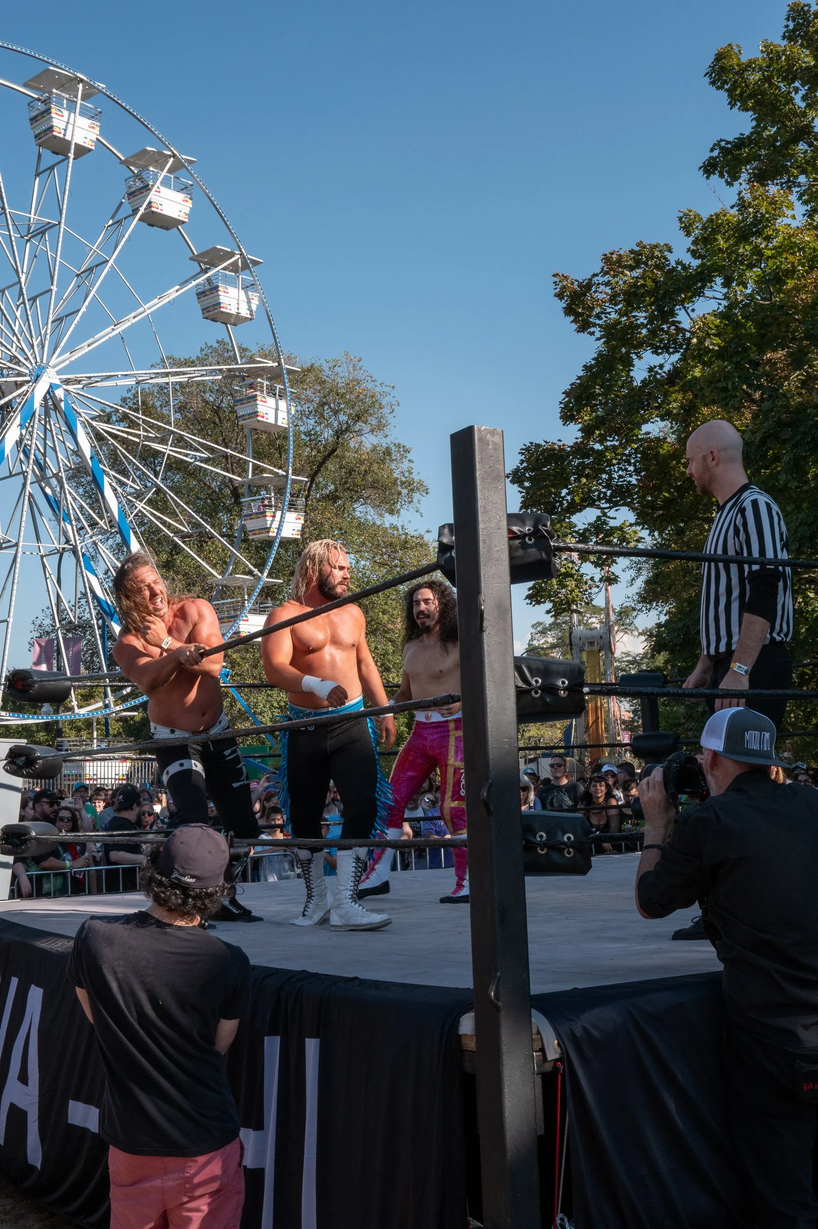 Wrestlers in colorful attire fighting in ring at outdoor amusement park with Ferris wheel and crowd watching on a sunny day.