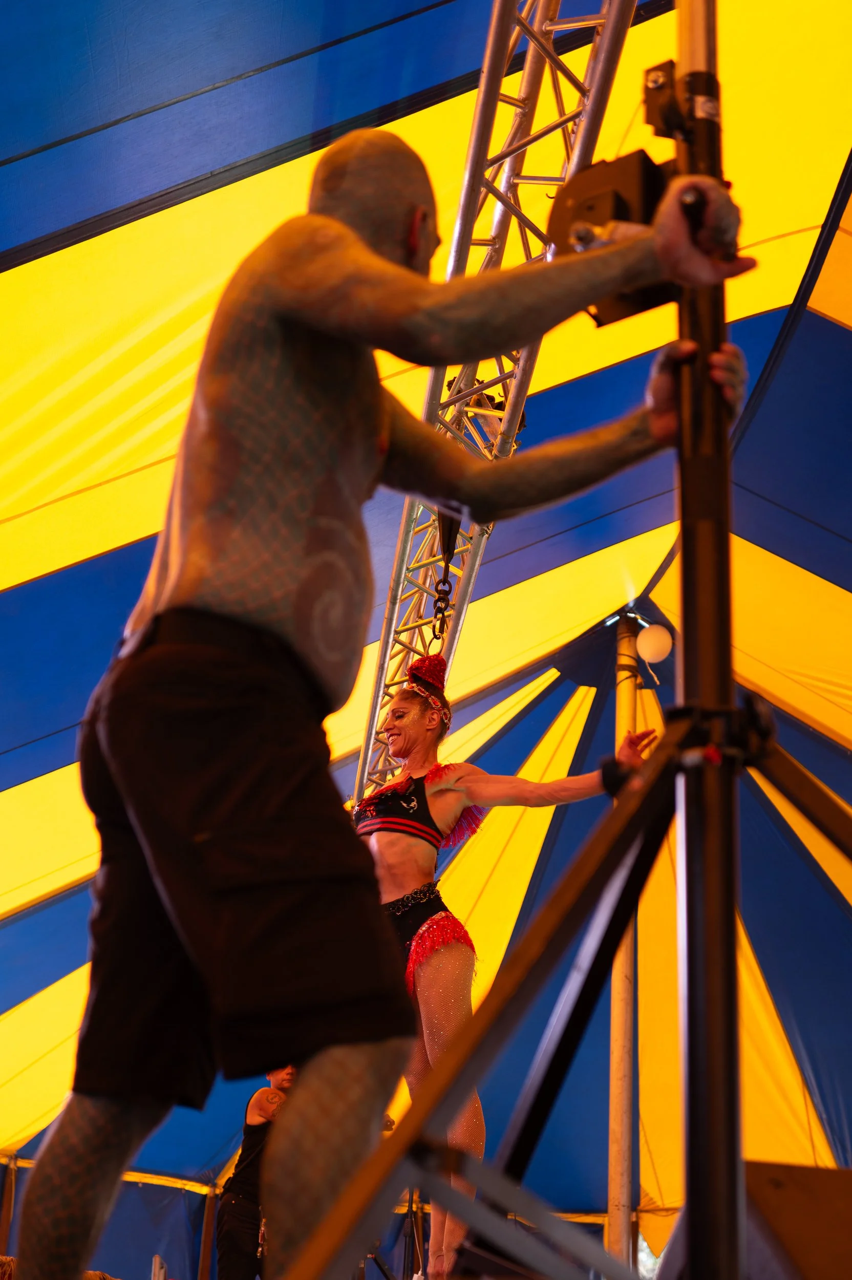 A performer in a red and black costume with a feathered headpiece is on a stage inside a yellow and blue circus tent, smiling and posing with her arms extended.