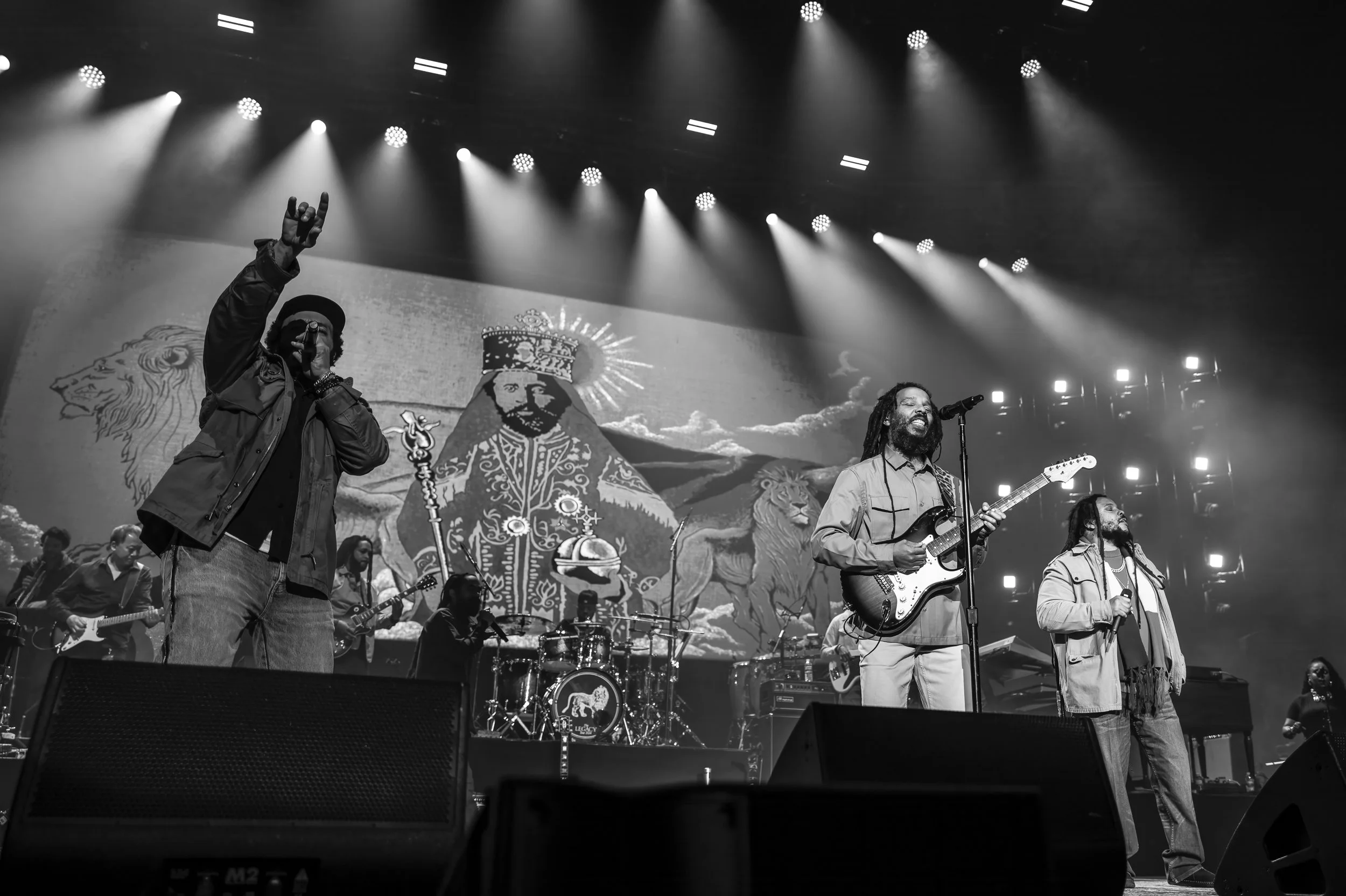 A black-and-white photo of a band performing on stage, featuring two vocalists in the foreground, with one raising their hand, and a guitarist on the right singing into a microphone. The background shows a large screen with artwork of a crowned figur