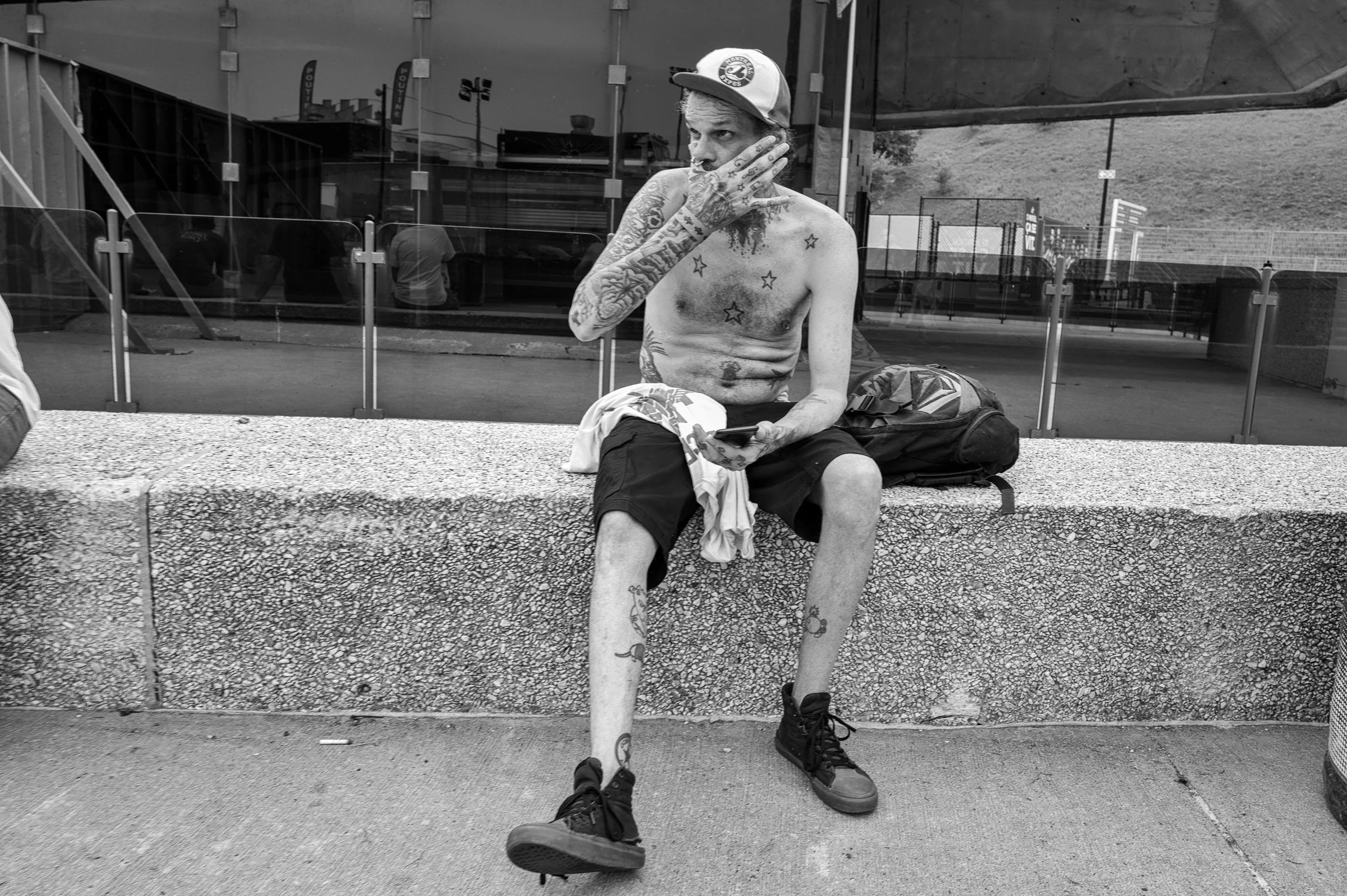 A shirtless man with numerous tattoos, wearing a baseball cap, sitting on a concrete bench at an outdoor sports facility, holding a phone in one hand and resting his head on his other hand.