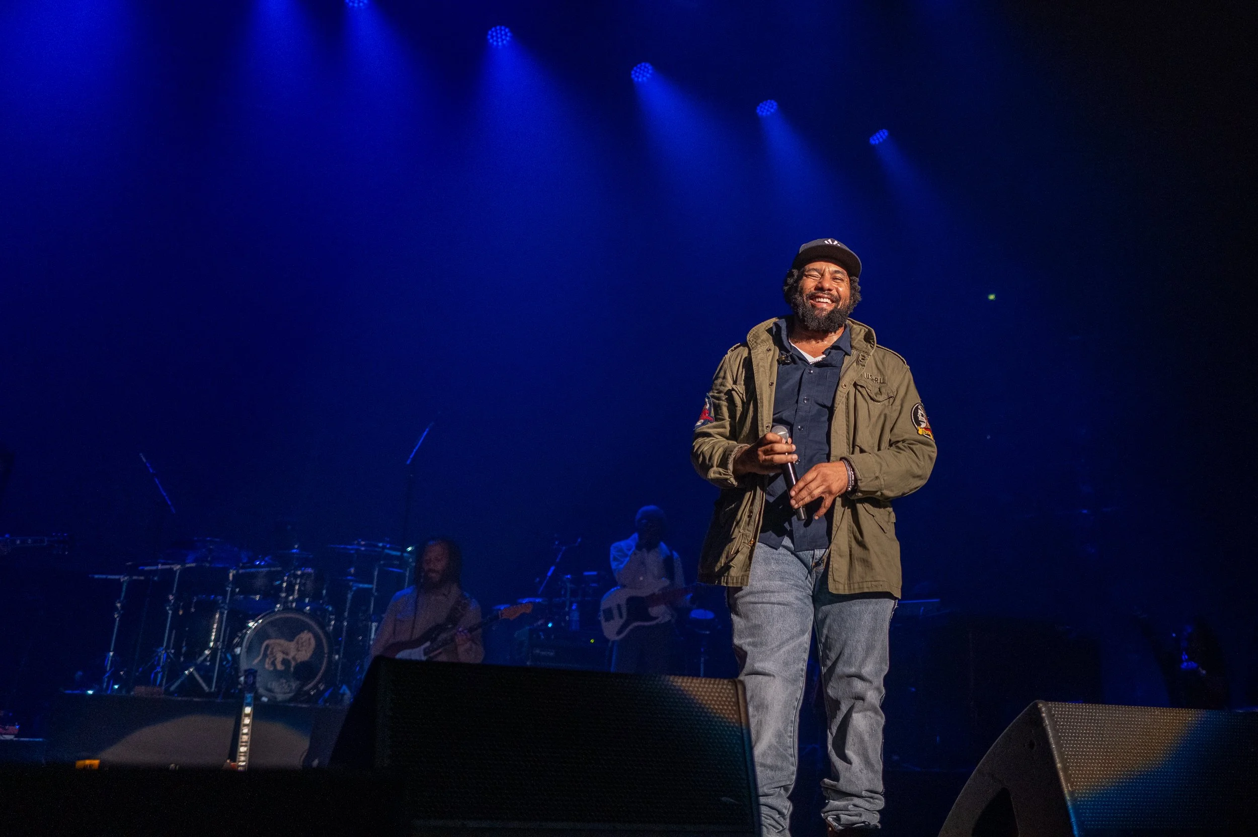 A man with a beard, wearing a dark cap and jacket, smiles while standing on stage with a microphone in hand, under blue stage lights, with band members in the background.