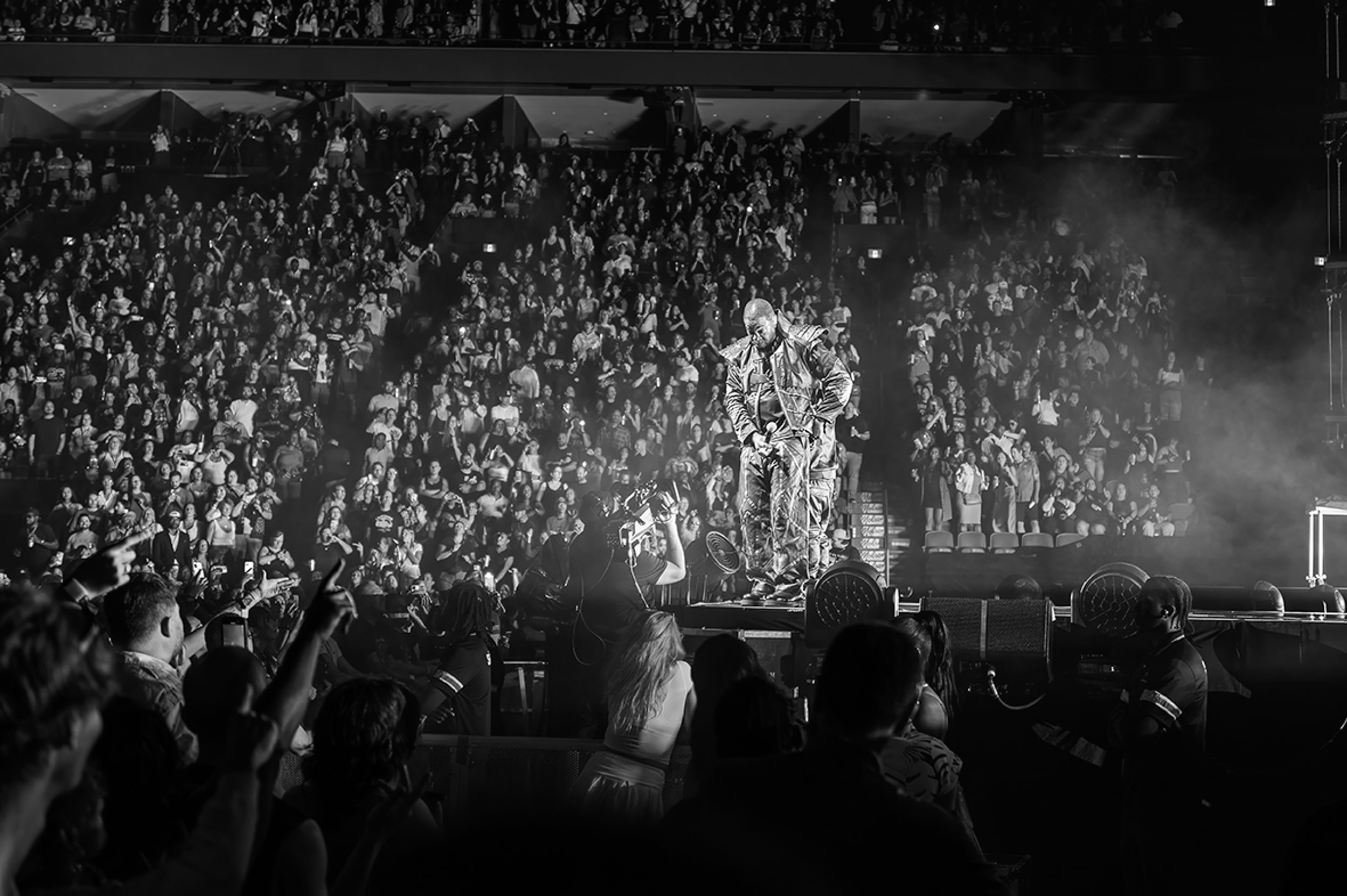 Black and white photo of a large indoor concert with a dense crowd. A performer in a shiny outfit is on stage, surrounded by a foggy atmosphere, with audience members taking photos and raising their hands.