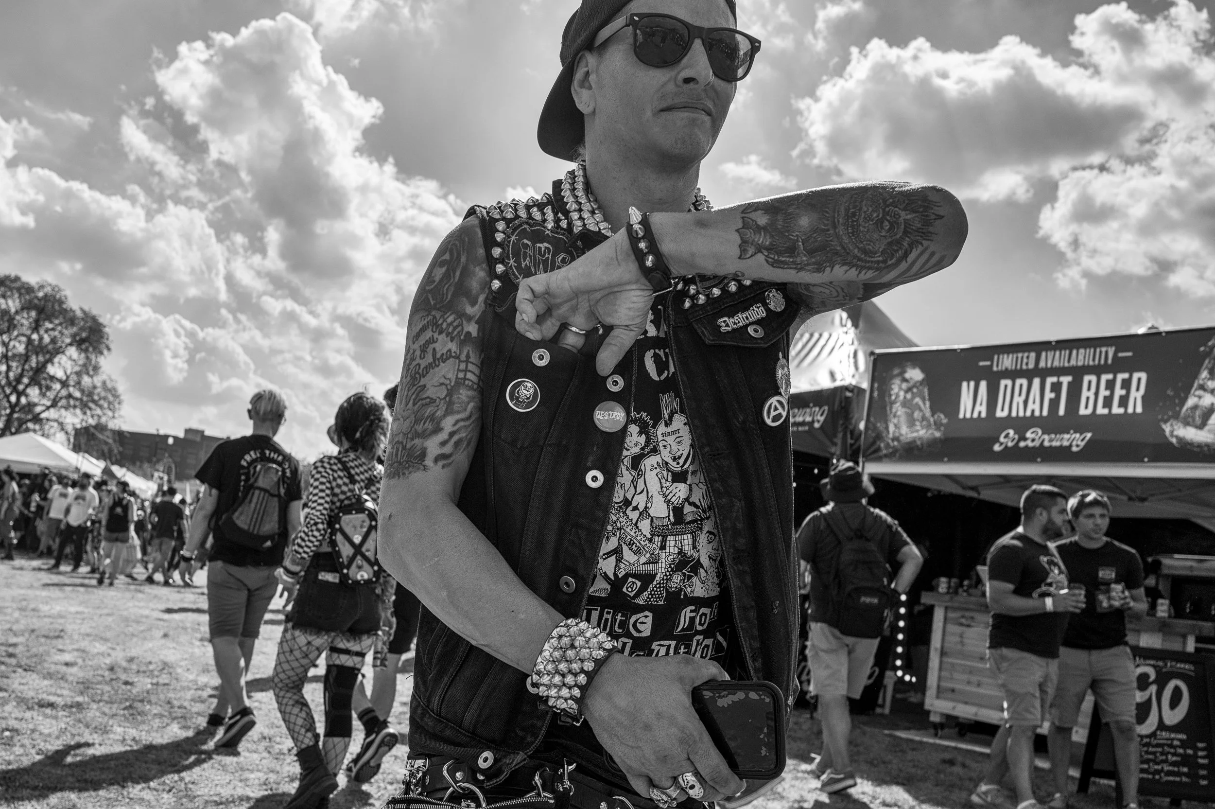 A man with tattoos, wearing sunglasses, a backward cap, and a studded vest, is flexing his arm in front of a crowd at an outdoor event. The scene is in black and white with a cloudy sky and vendor booths in the background.
