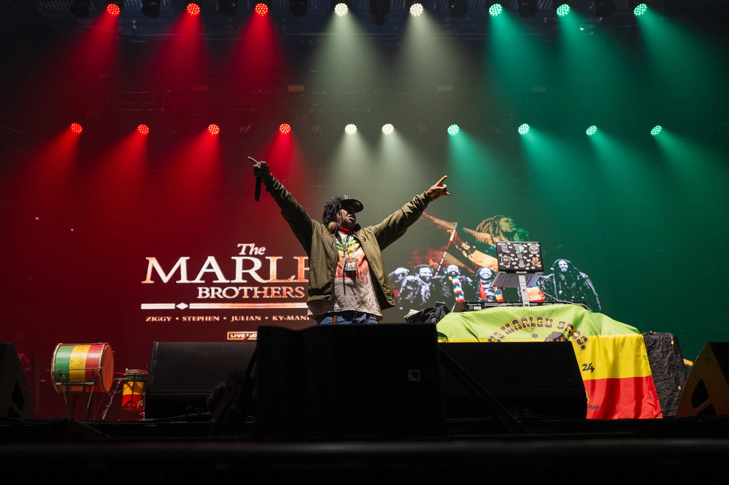 A DJ performs on stage with arms raised, wearing headphones and a jacket. Behind him, a screen displays 'The MARLEY BROTHERS' with images of Bob Marley and others. The stage has reggae-themed decorations, including a drum with reggae colors and a tab