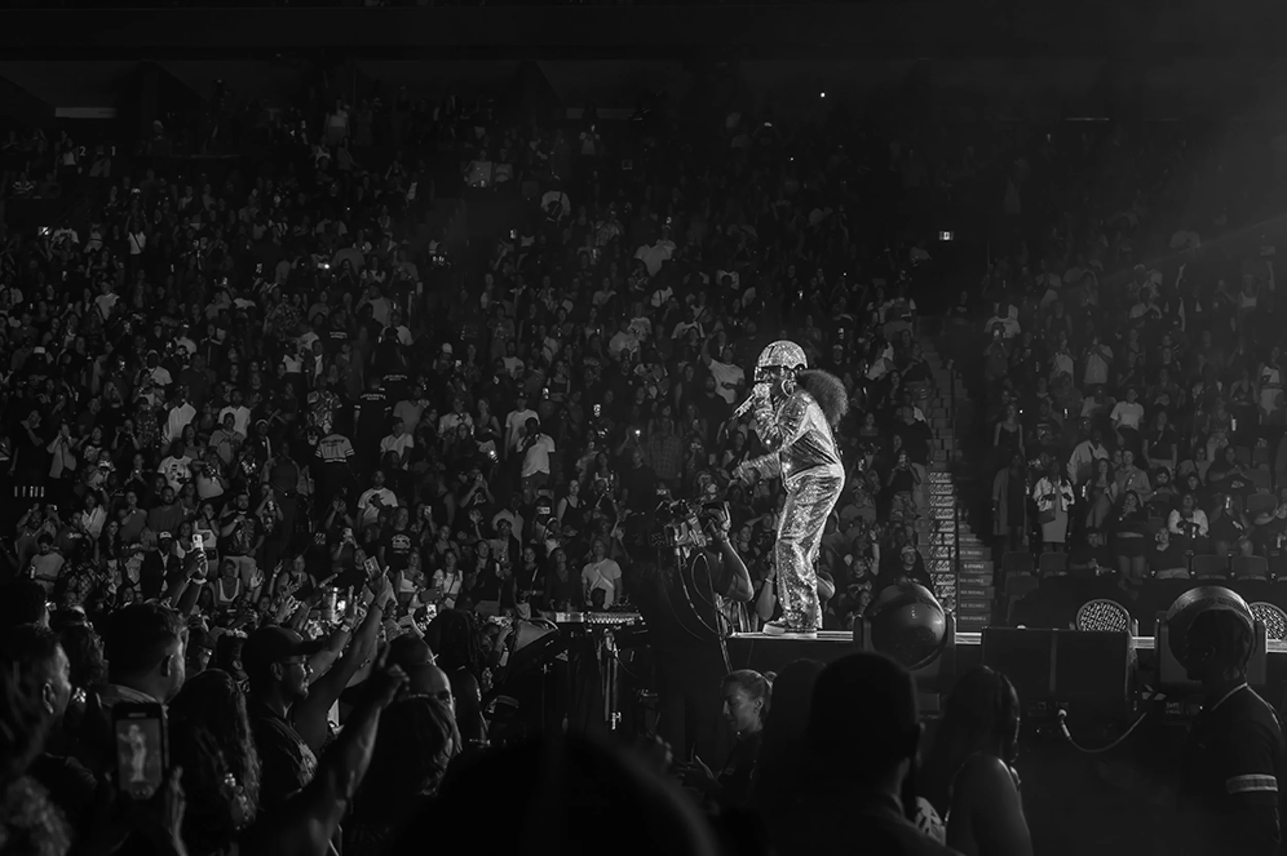 Performing artist on stage with a microphone in front of a large, indoor audience, wearing shiny clothing and a head covering. Audience members are capturing the moment with phones.