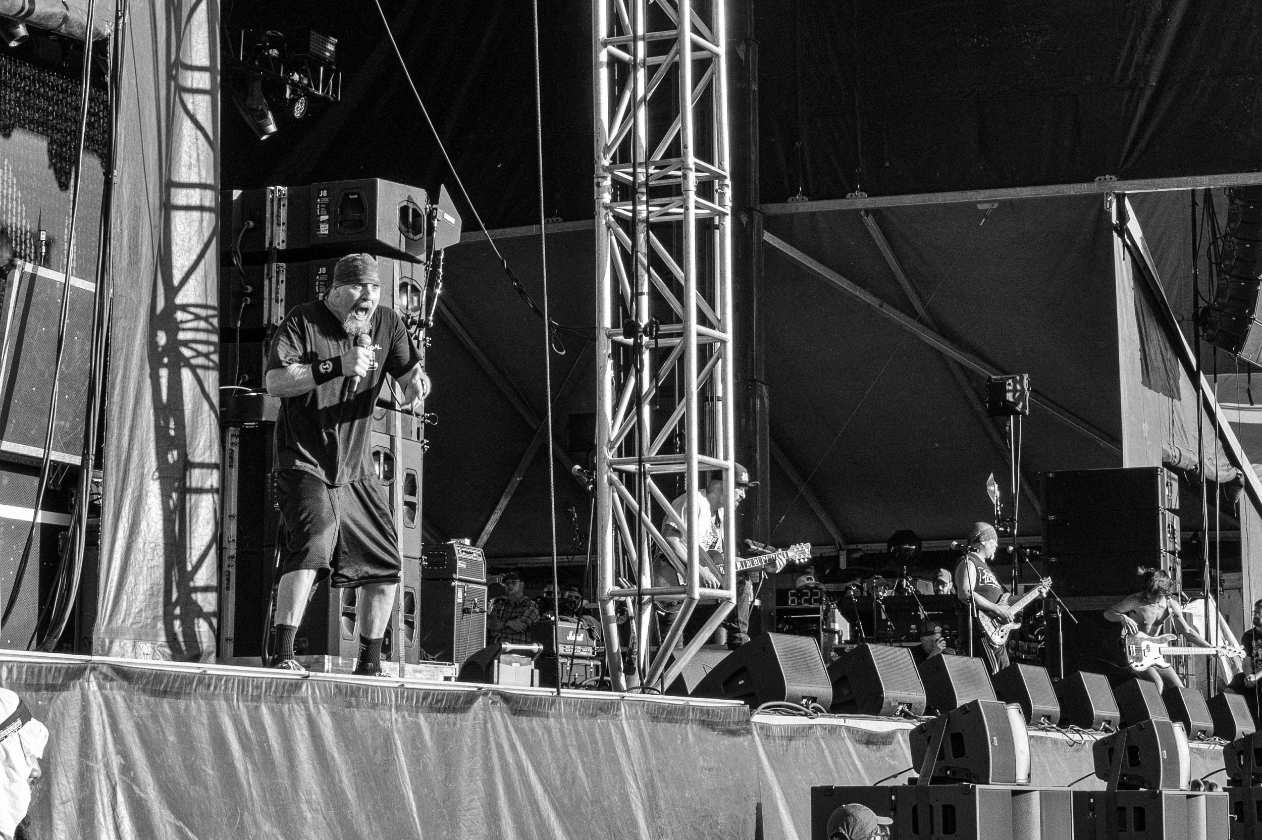 Black and white photo of a band performing on stage at a concert, with a singer holding a microphone and three guitarists playing in the background.