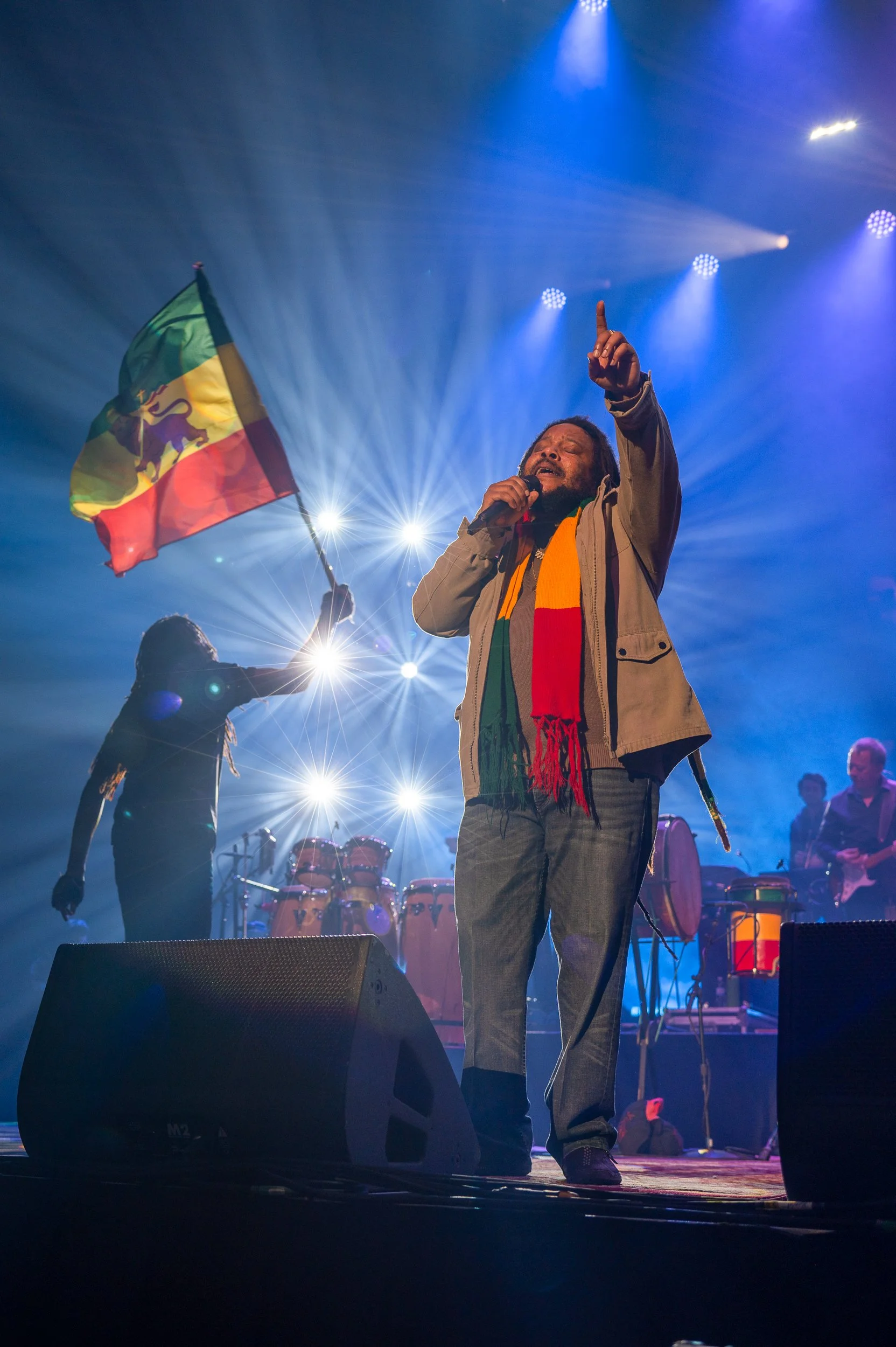 A man singing on stage with a microphone, wearing a scarf with colors of the Ethiopian flag, and holding an Ethiopian flag. Bright stage lights create a dramatic backdrop.