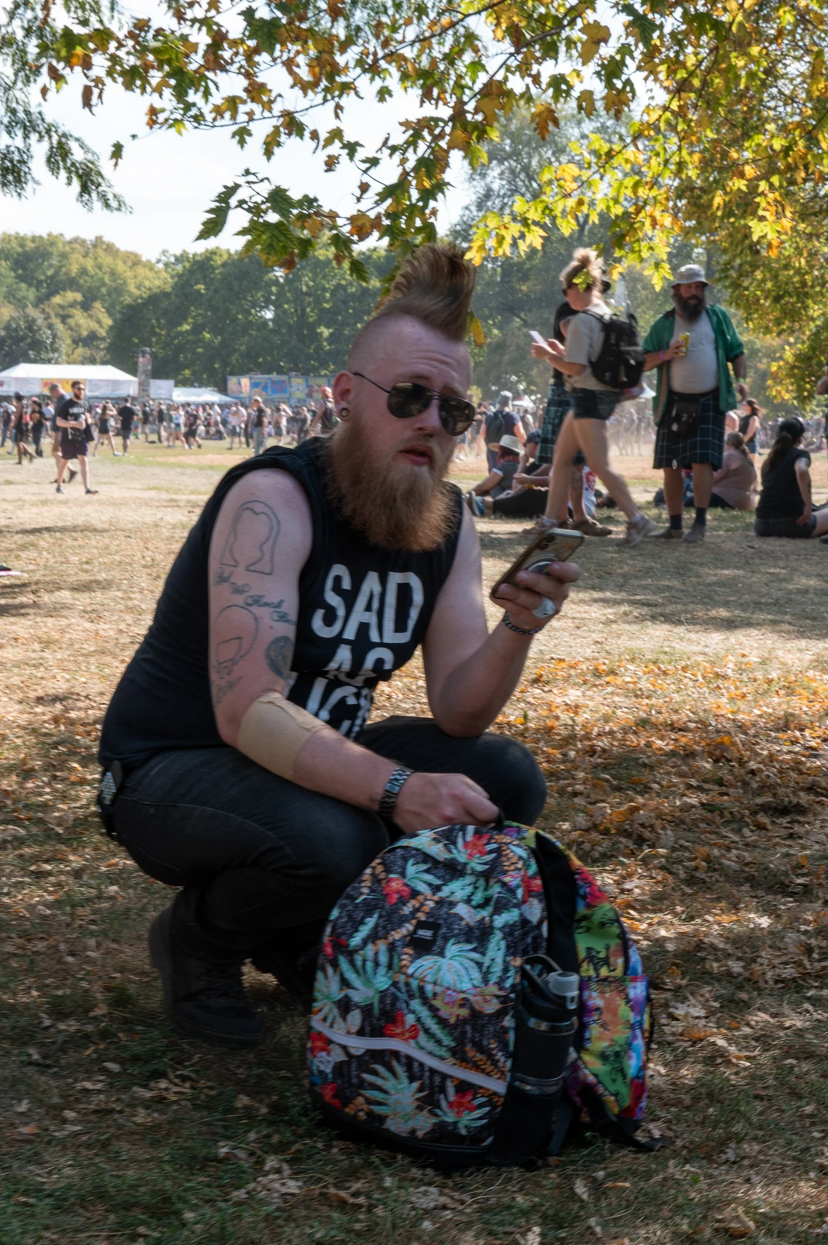 A man with tattoos, sunglasses, and a Mohawk hairstyle crouches on the grass at an outdoor festival, holding a phone. A colorful backpack is on the ground beside him, and there are other festival-goers in the background under a tree canopy.