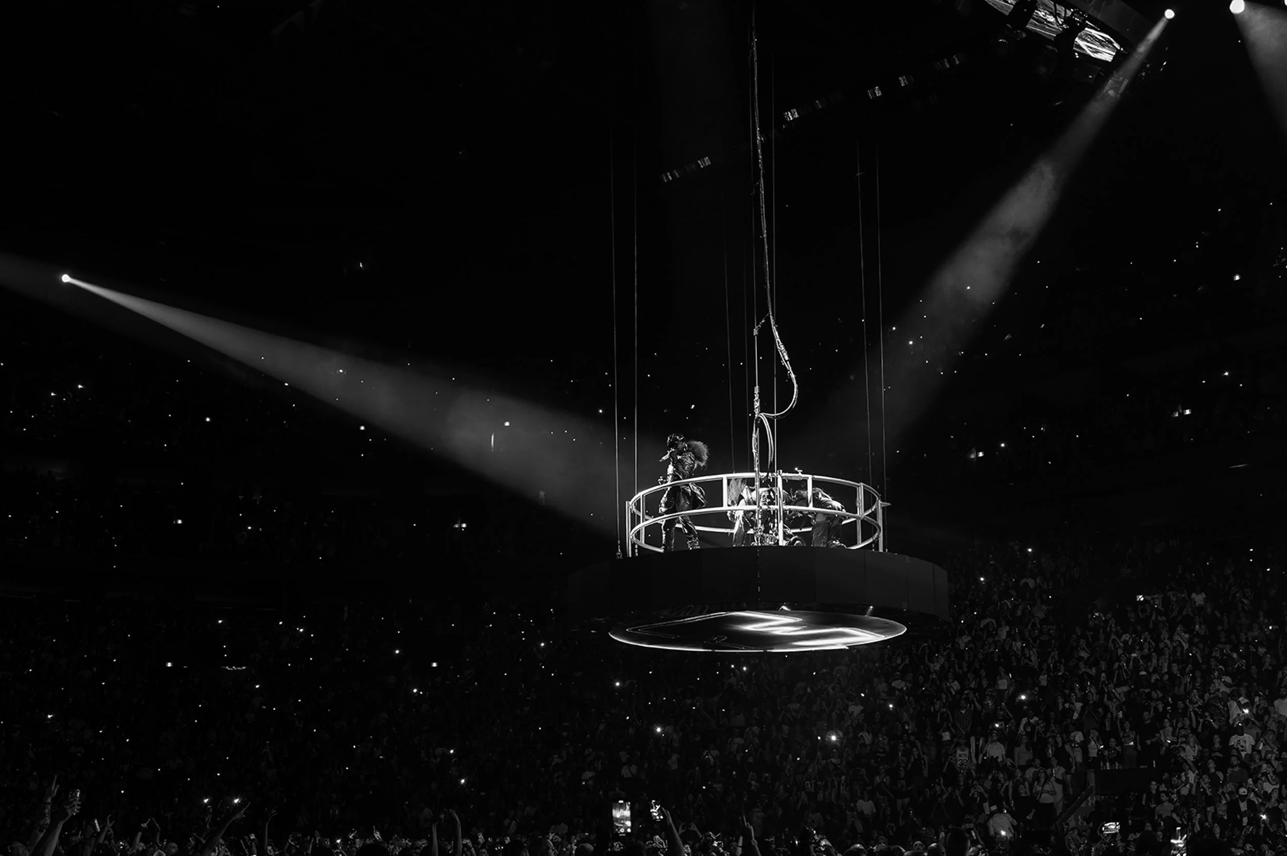 A concert scene with a performer on a suspended circular platform, illuminated by spotlights, with a large crowd in the background.