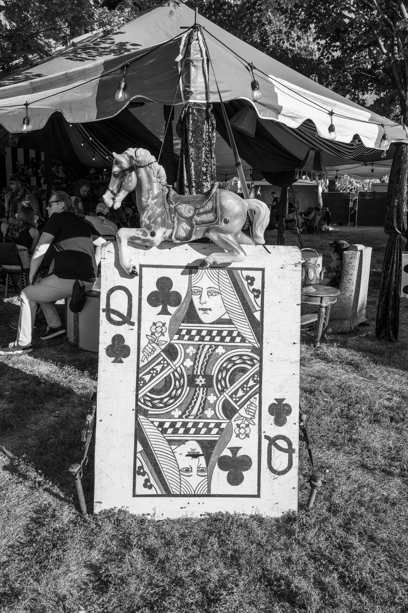Black and white photo of a large playing card decoration with the Queen of Clubs on a grassy area. A vintage rocking horse is on top of the card, and a carnival tent with people inside is in the background.