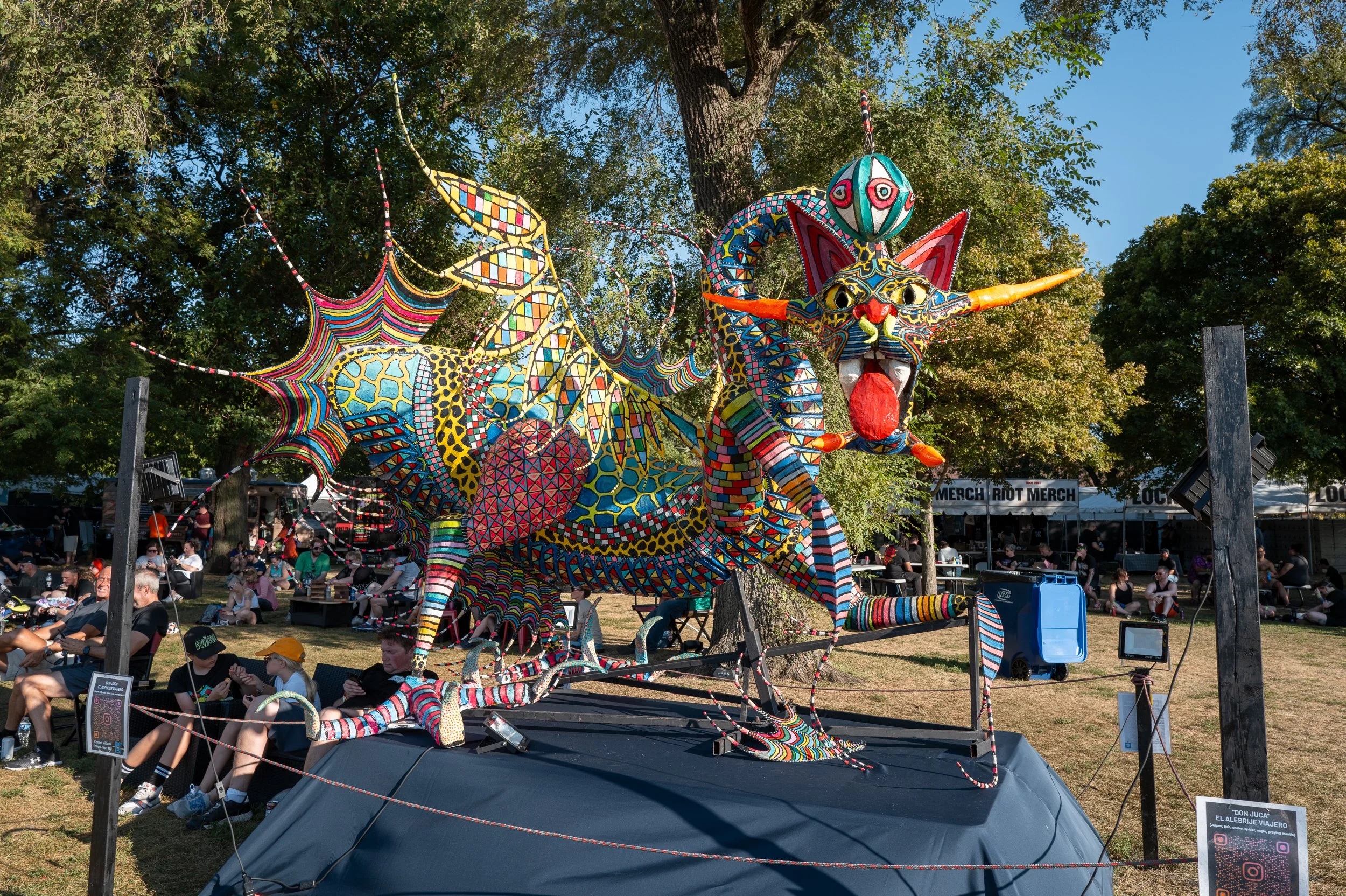Colorful dragon sculpture with intricate patterns, displayed outdoors, surrounded by trees and people sitting and walking in the background.