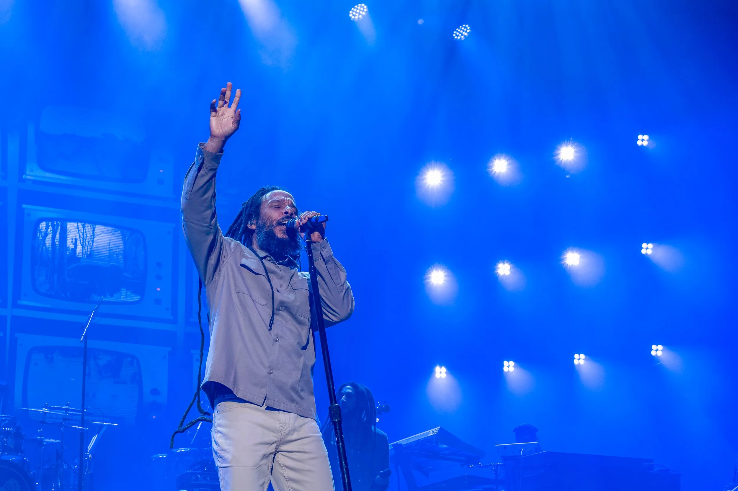 Male singer with dreadlocks and beard, wearing beige shirt and pants, singing into microphone with eyes closed on stage illuminated by blue and white lights.