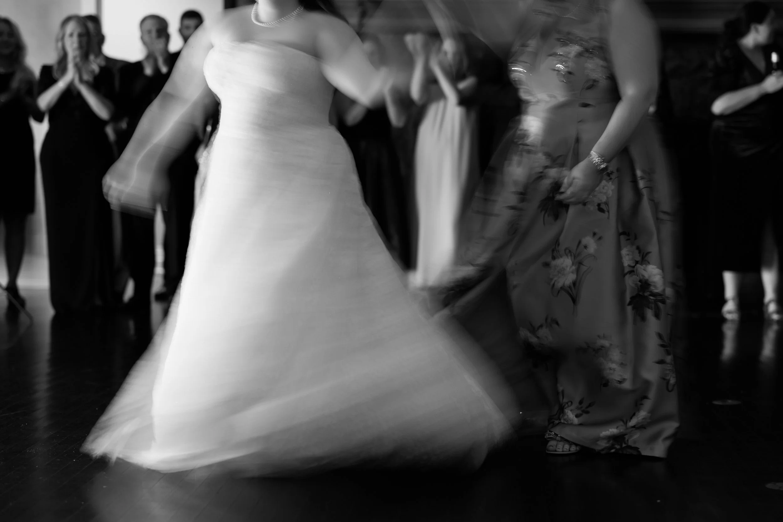 A woman in a wedding dress is dancing with a woman in a floral dress at a wedding reception, surrounded by guests.