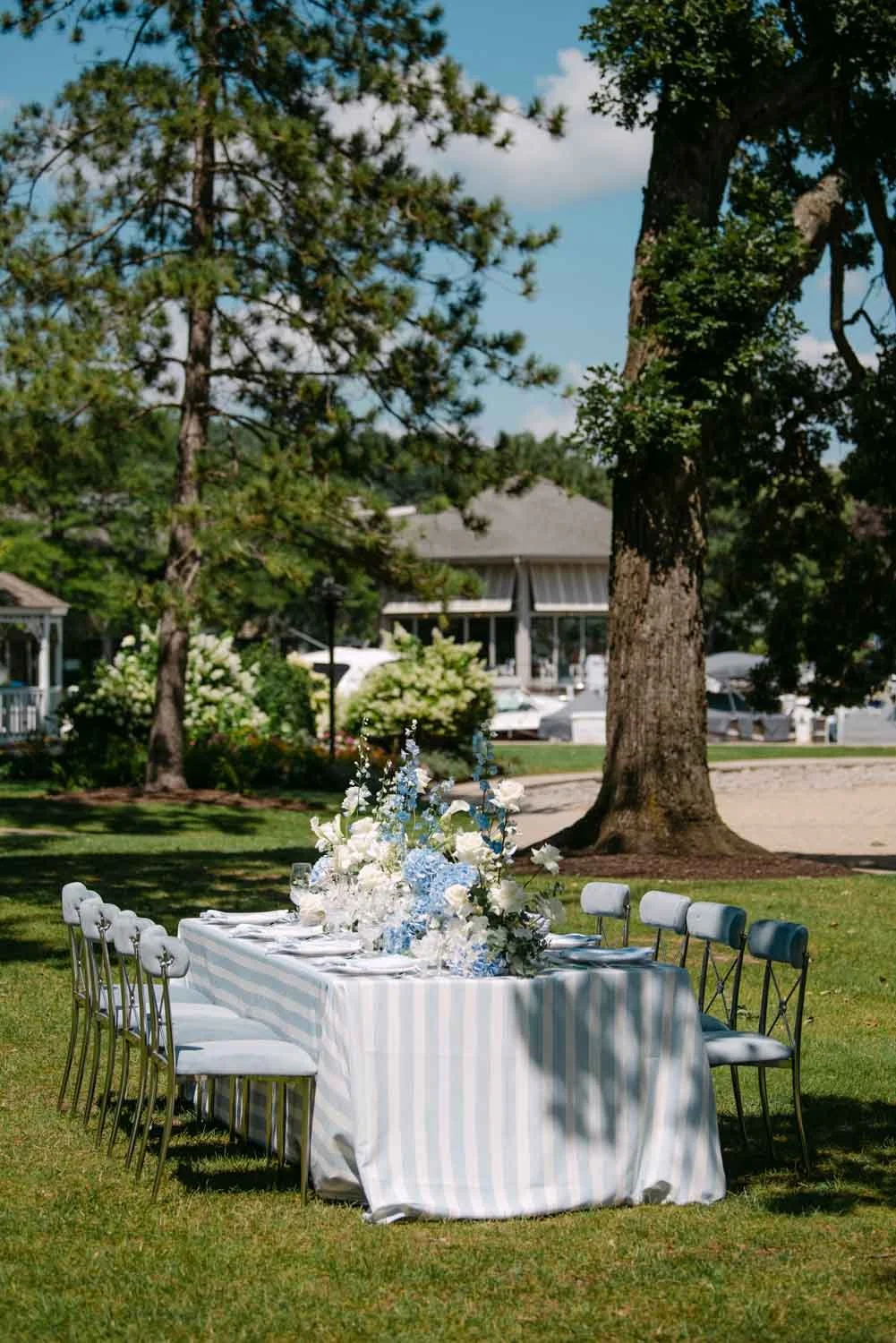 Outdoor banquet table decorated with a striped tablecloth and floral centerpieces in a park-like setting with trees and boats in the background.