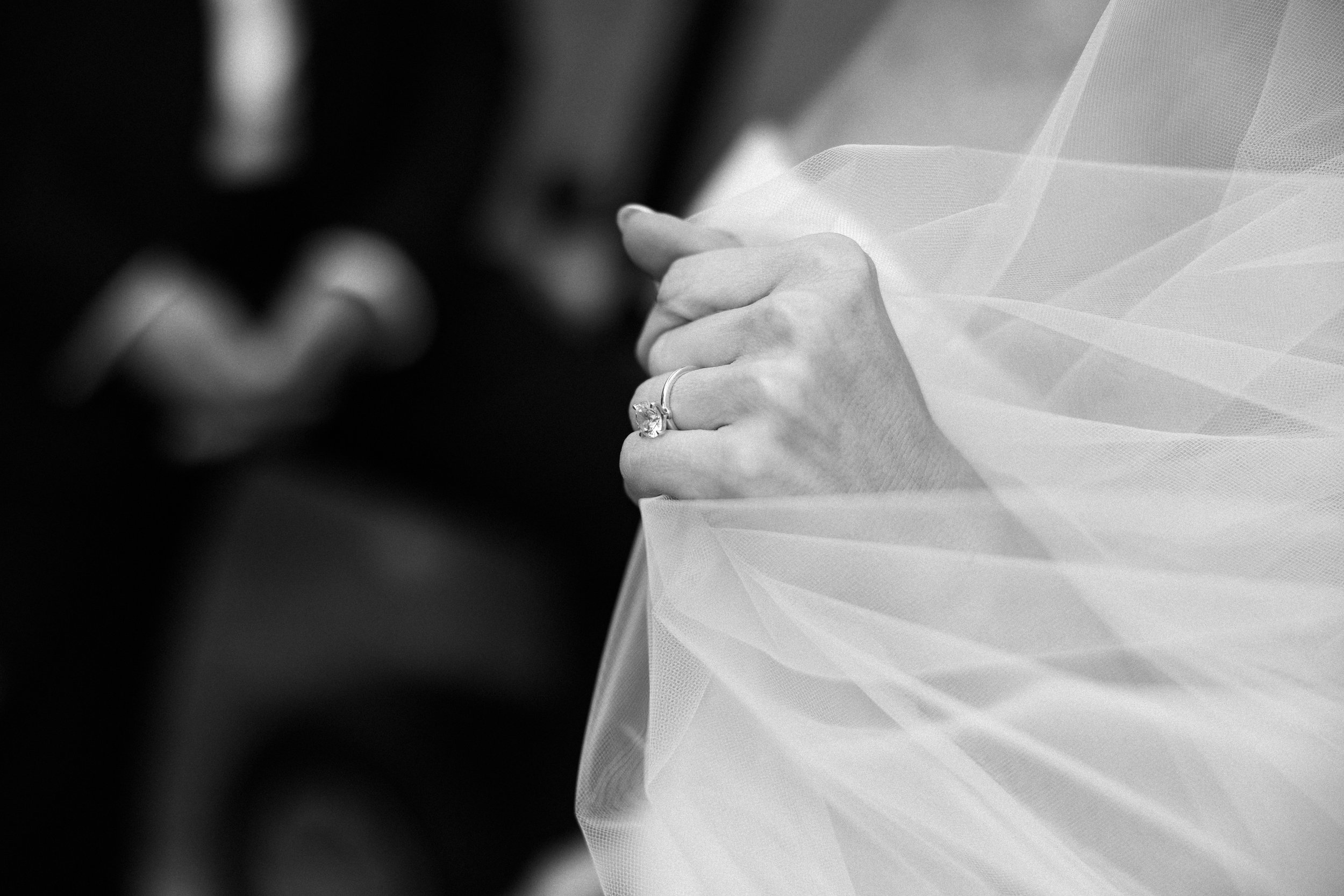 Close-up of a hand with an engagement ring, holding the edge of a wedding dress, in black and white.