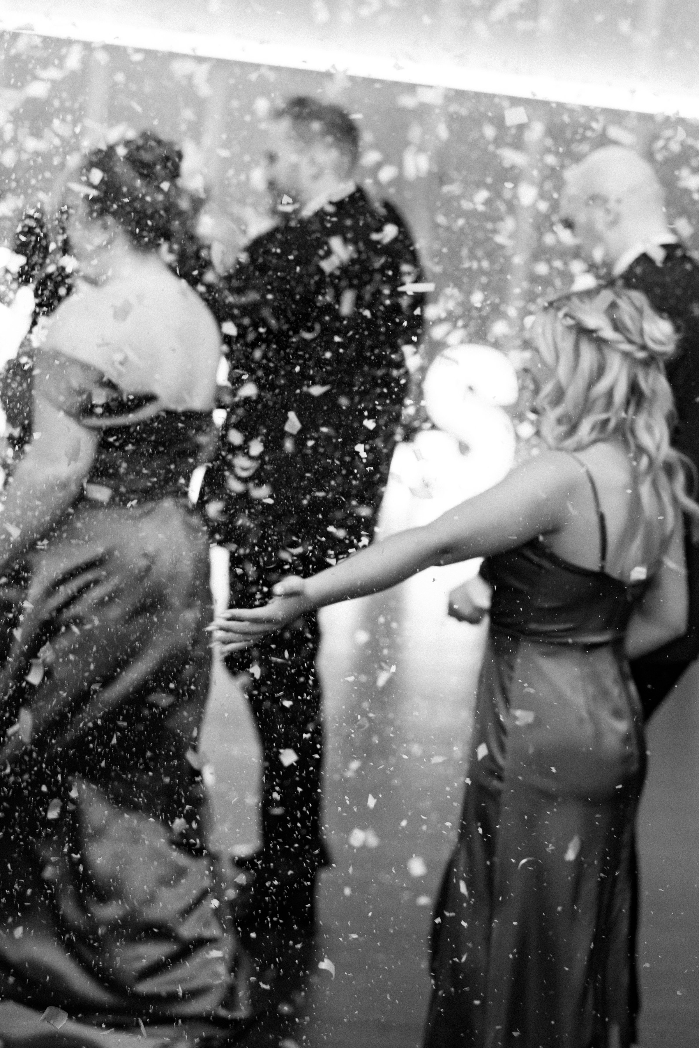 A woman in a slip dress reaching out to a man in a tuxedo amidst falling confetti at what appears to be a celebration or event.