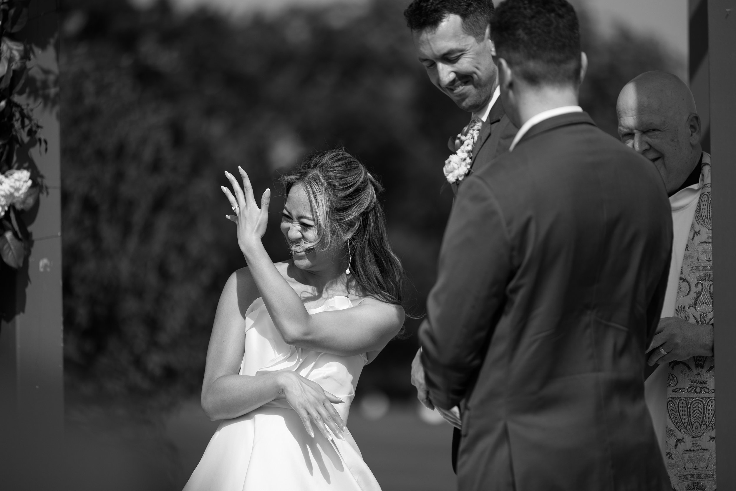 Black and white photo of a woman smiling and showing her wedding ring, surrounded by two men in suits and an officiant during a wedding ceremony outdoors.