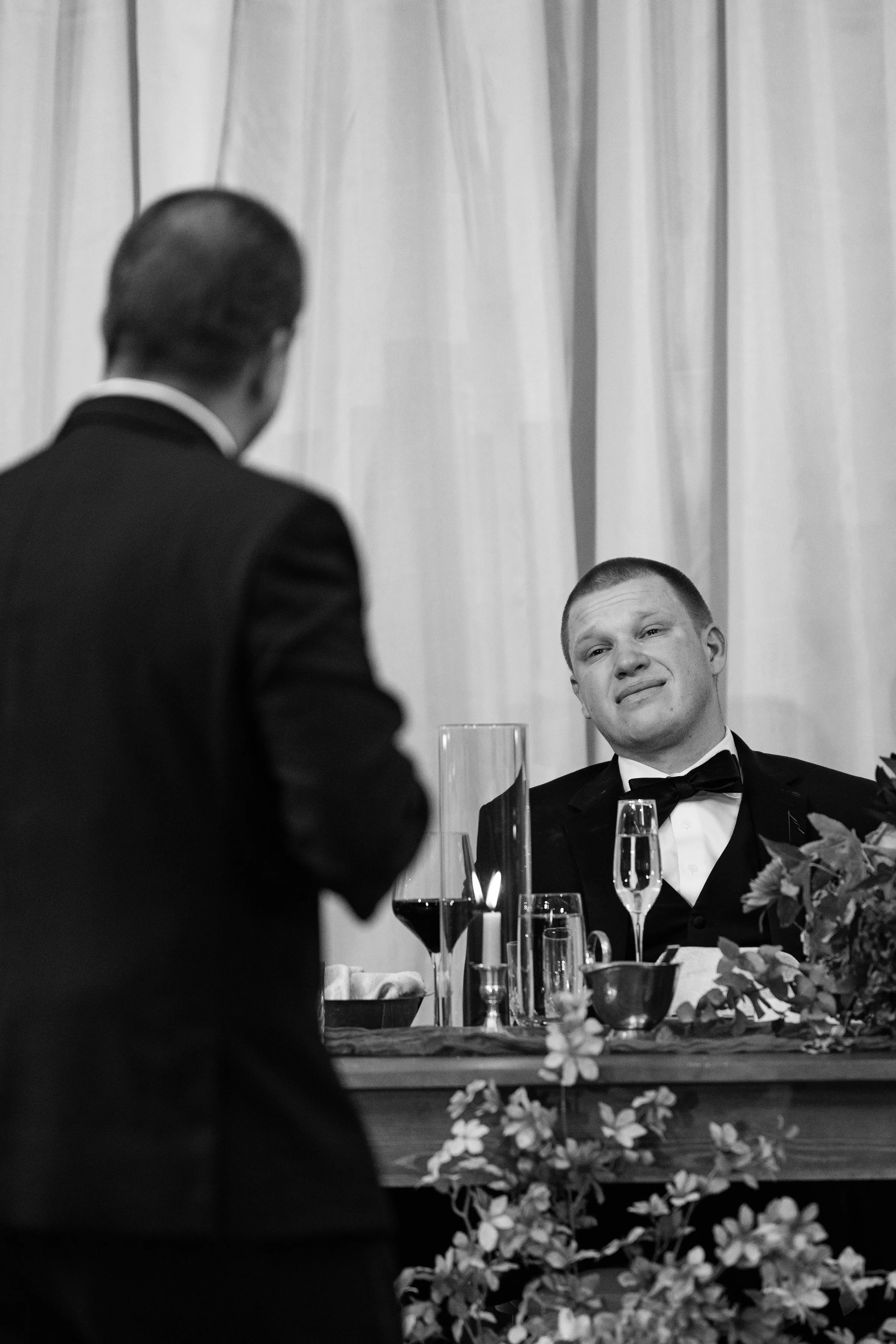A black and white photograph of a man in a tuxedo with a bow tie sitting at a decorated table with flowers, wine, and champagne glasses. He appears to be listening and smirking slightly while another man, with his back turned and out of focus, appear