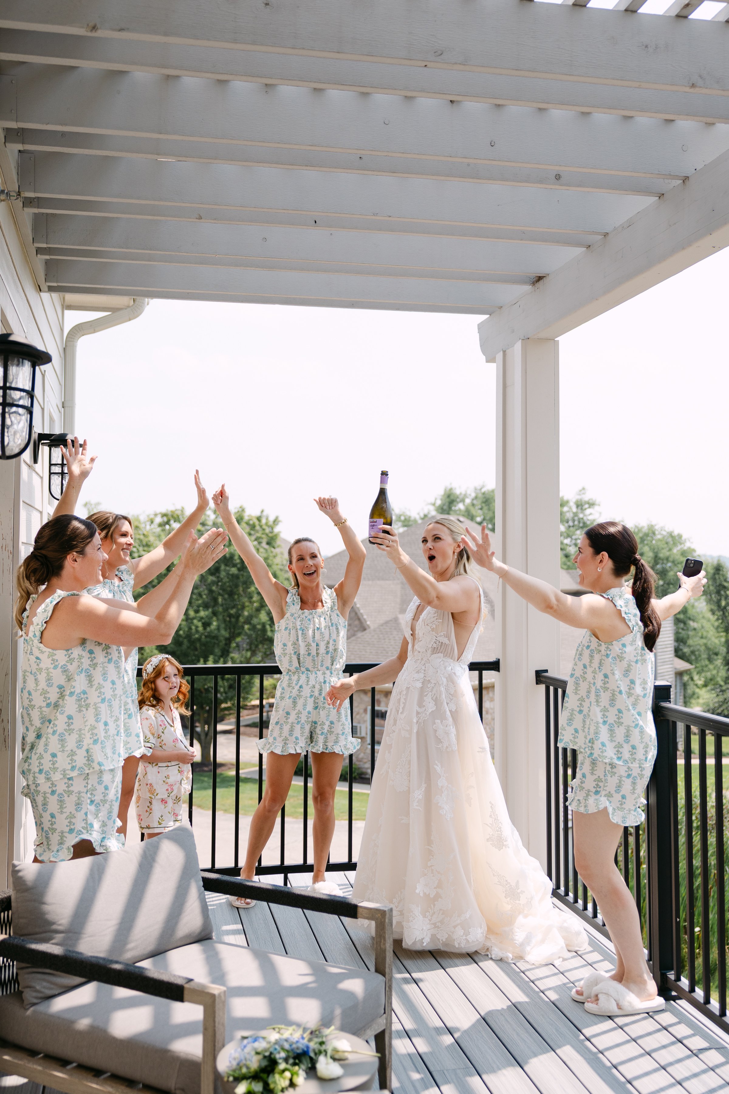 A woman in a wedding dress holding a bottle of champagne on a balcony surrounded by her friends in pajamas celebrating.