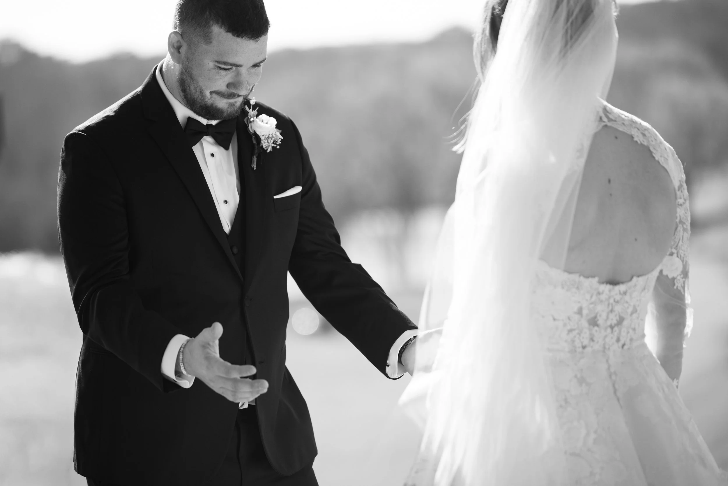 A groom in a black tuxedo and bow tie is holding the hand of a bride wearing a lace wedding gown and veil, during a wedding ceremony outdoors.