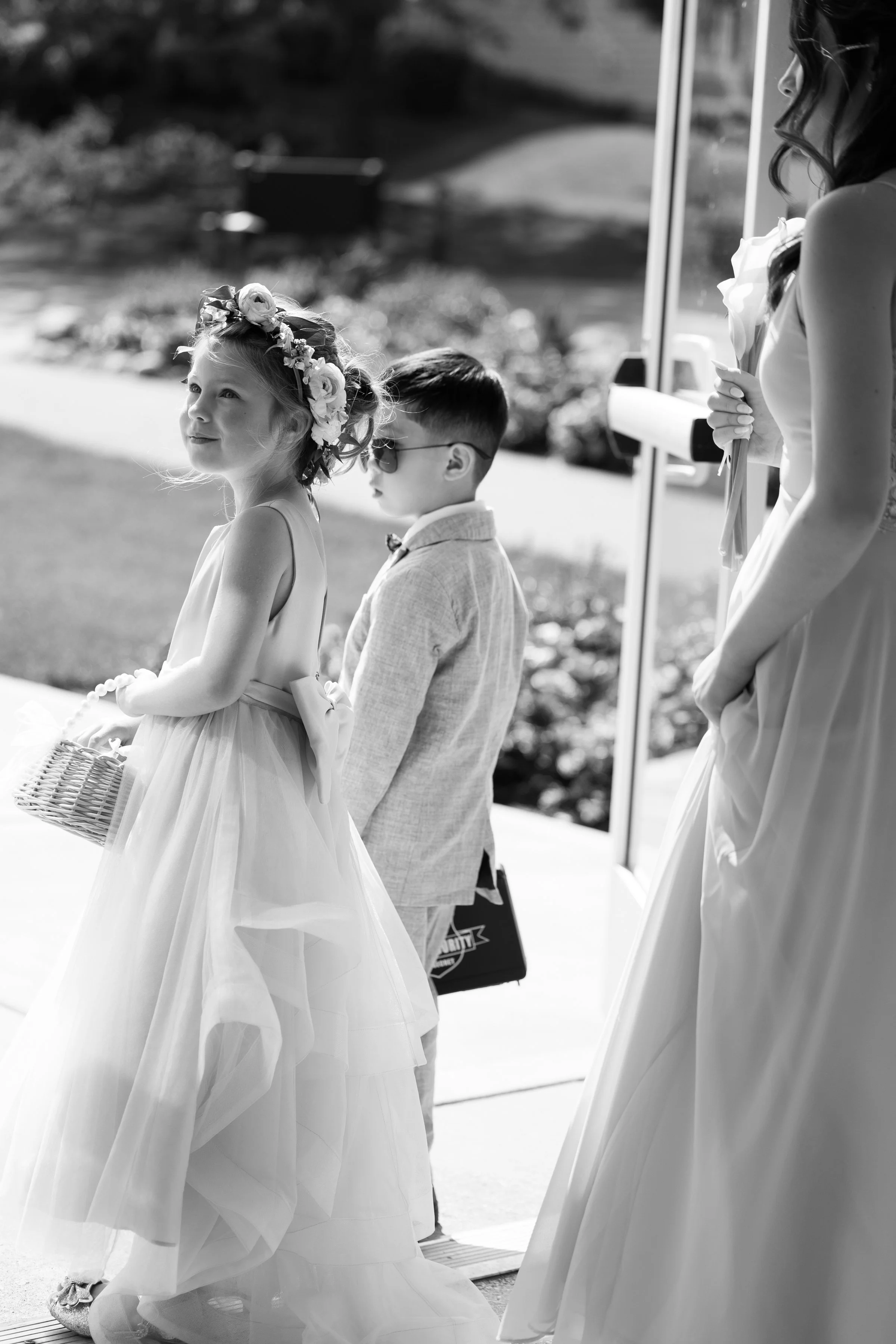 Young girl in a wedding dress wearing a floral crown, holding a basket, standing with a young boy in a light-colored suit and sunglasses, and a woman in a wedding dress holding a bouquet, at a wedding ceremony outdoors.