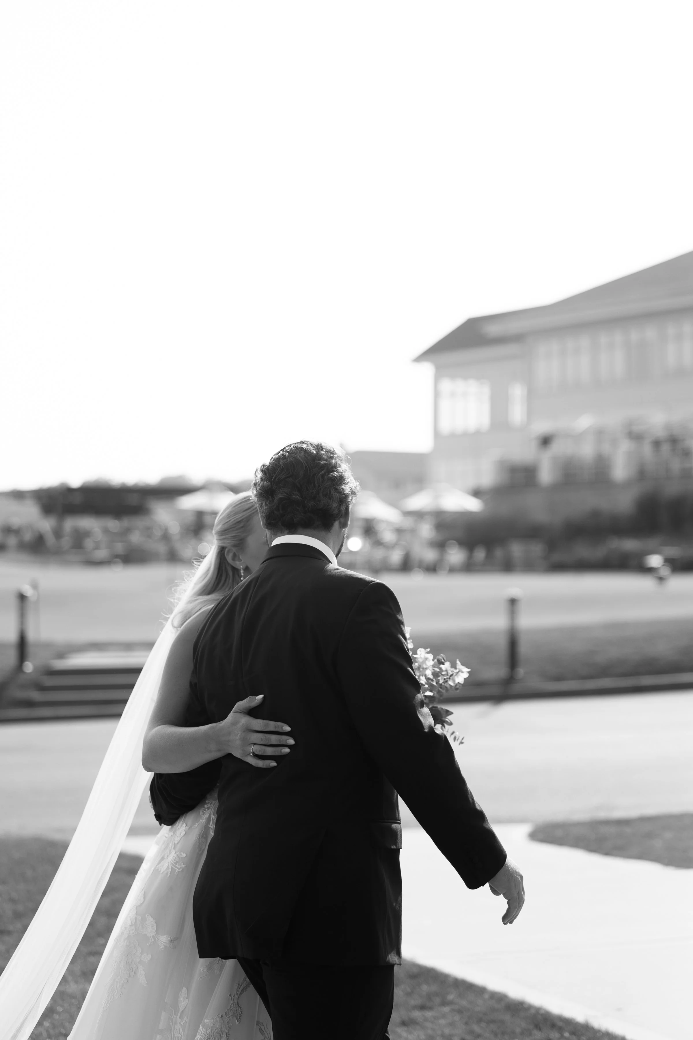 A bride and groom share an embrace outdoors, with the bride in a wedding dress and veil, and the groom in a suit, holding a bouquet of flowers.