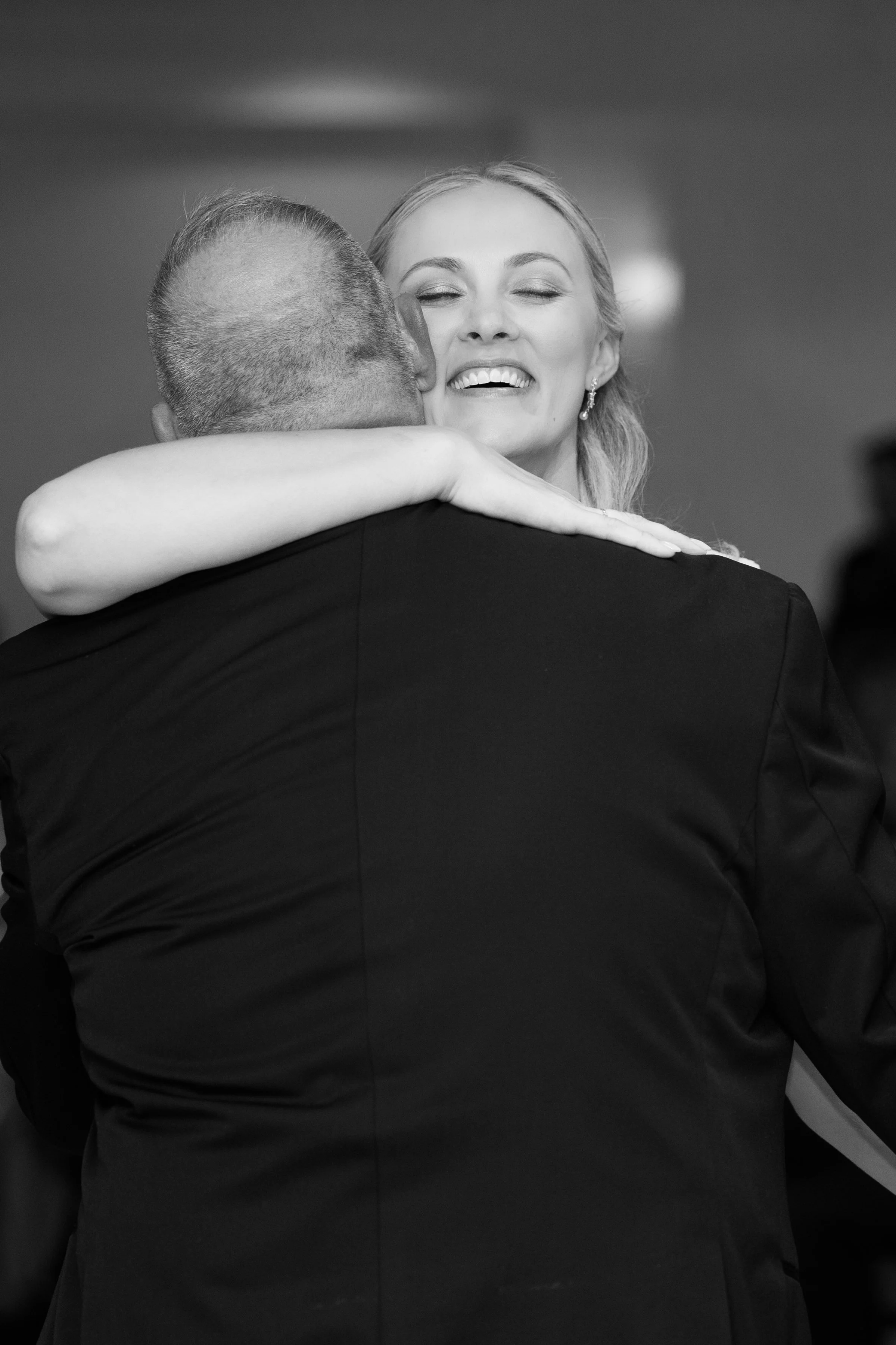 A woman with long hair smiling with her eyes closed, hugging a man in a suit from behind, in a black and white photo.