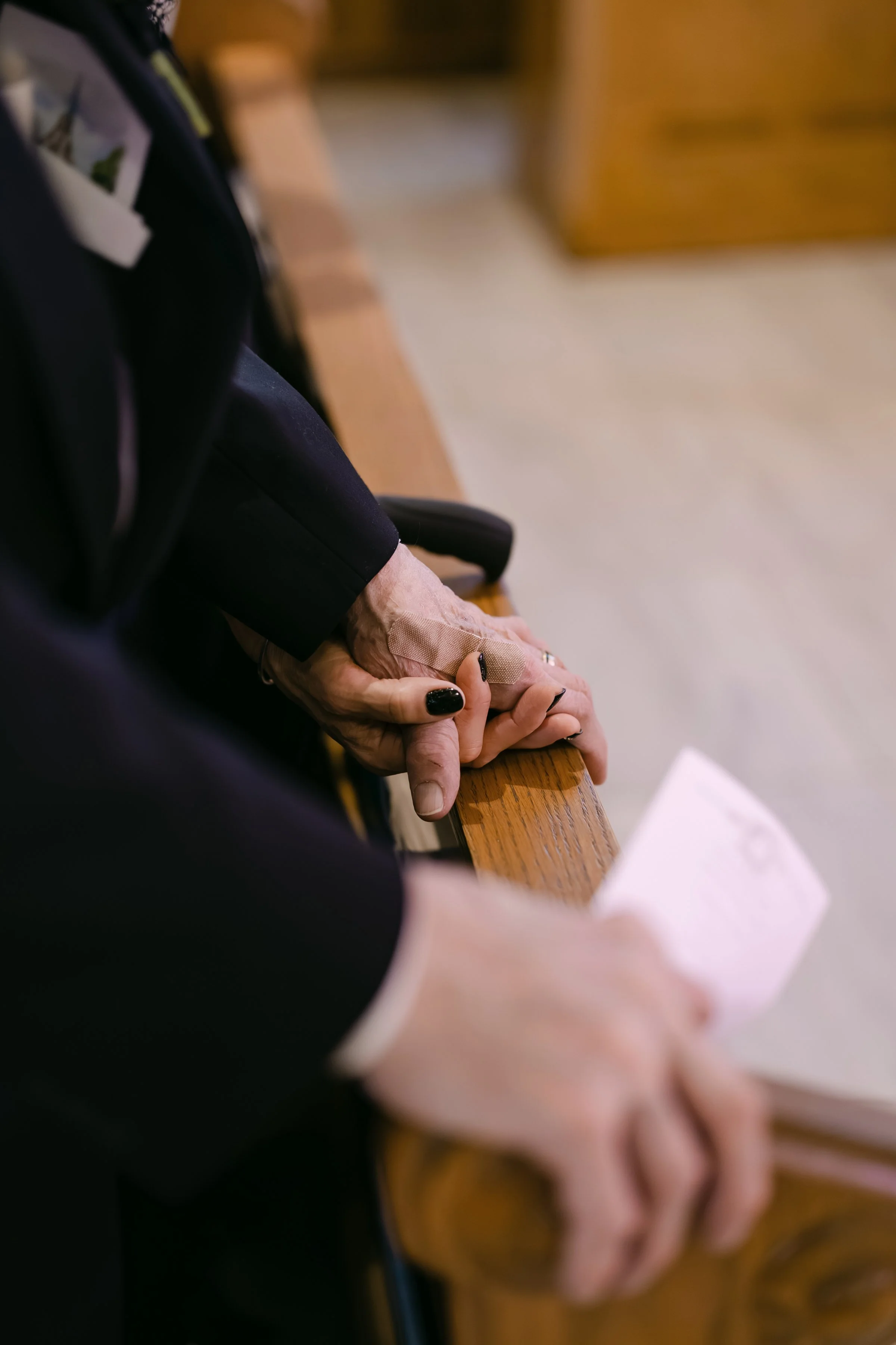 Close-up of an elderly person's clasped hands resting on a wooden pew inside a church or chapel.