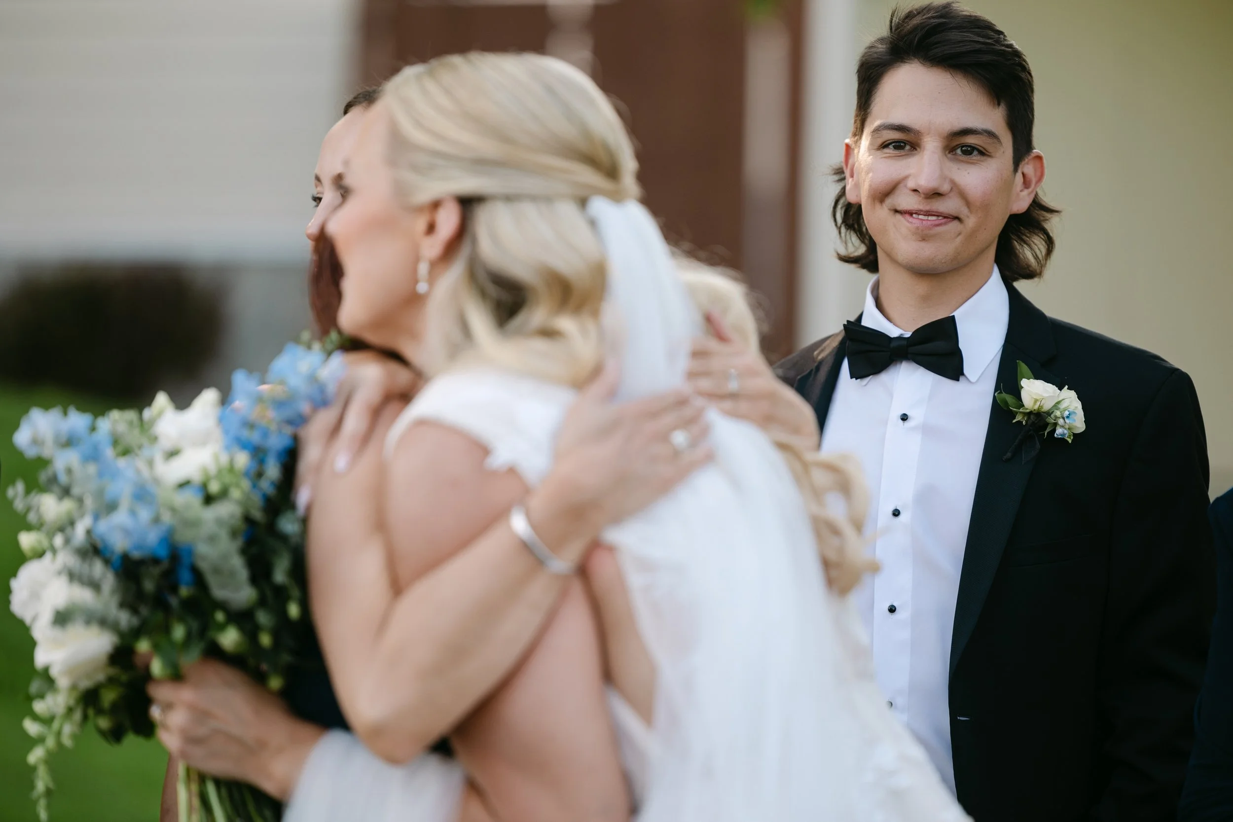 A wedding scene with a bride in a white gown and veil embracing a woman, while a smiling groom in a black tuxedo and bow tie watches. The bride holds a bouquet of blue and white flowers.