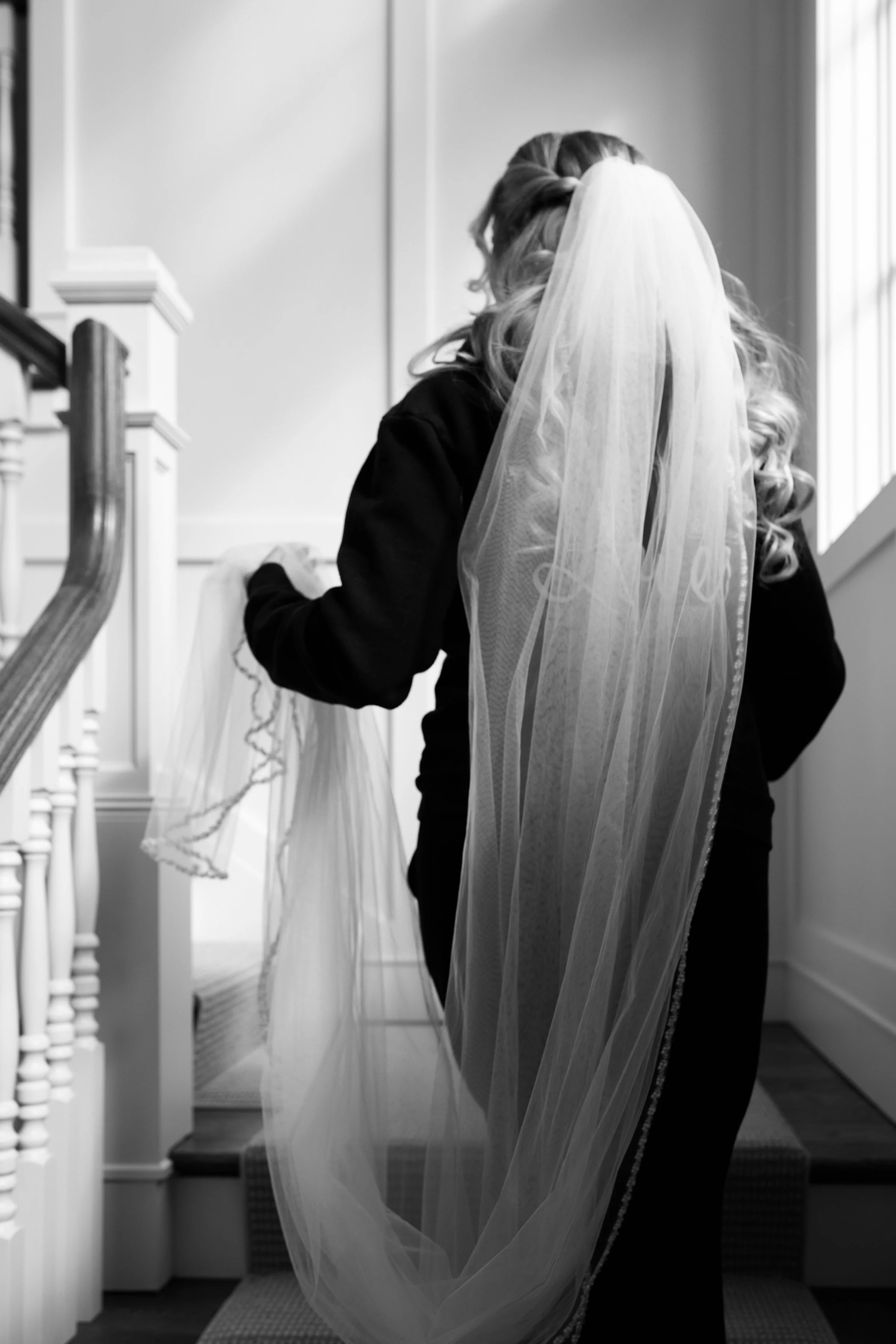 A woman with long, curly hair, facing away, holding a long wedding veil in a home interior with stairs and a large window.