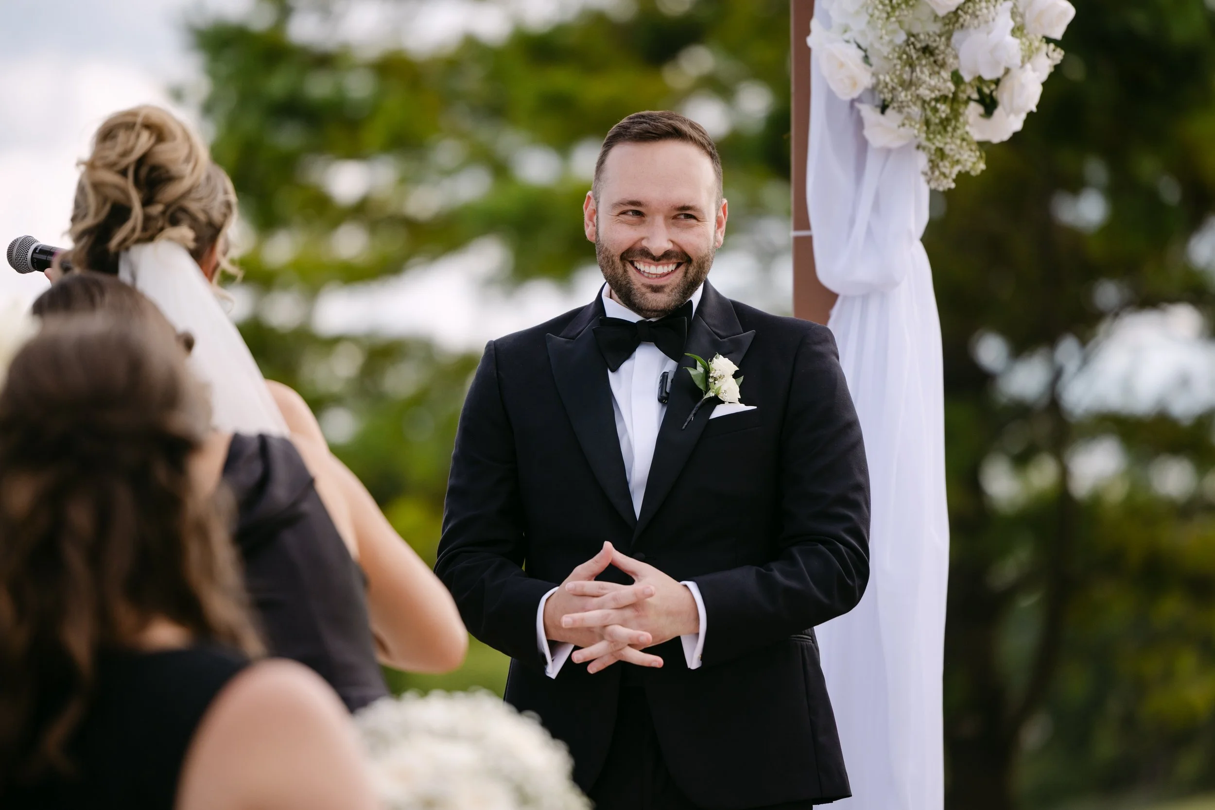 A groom in a black tuxedo with a boutonniere is smiling during a wedding ceremony outdoors, with a white floral arrangement and greenery in the background.