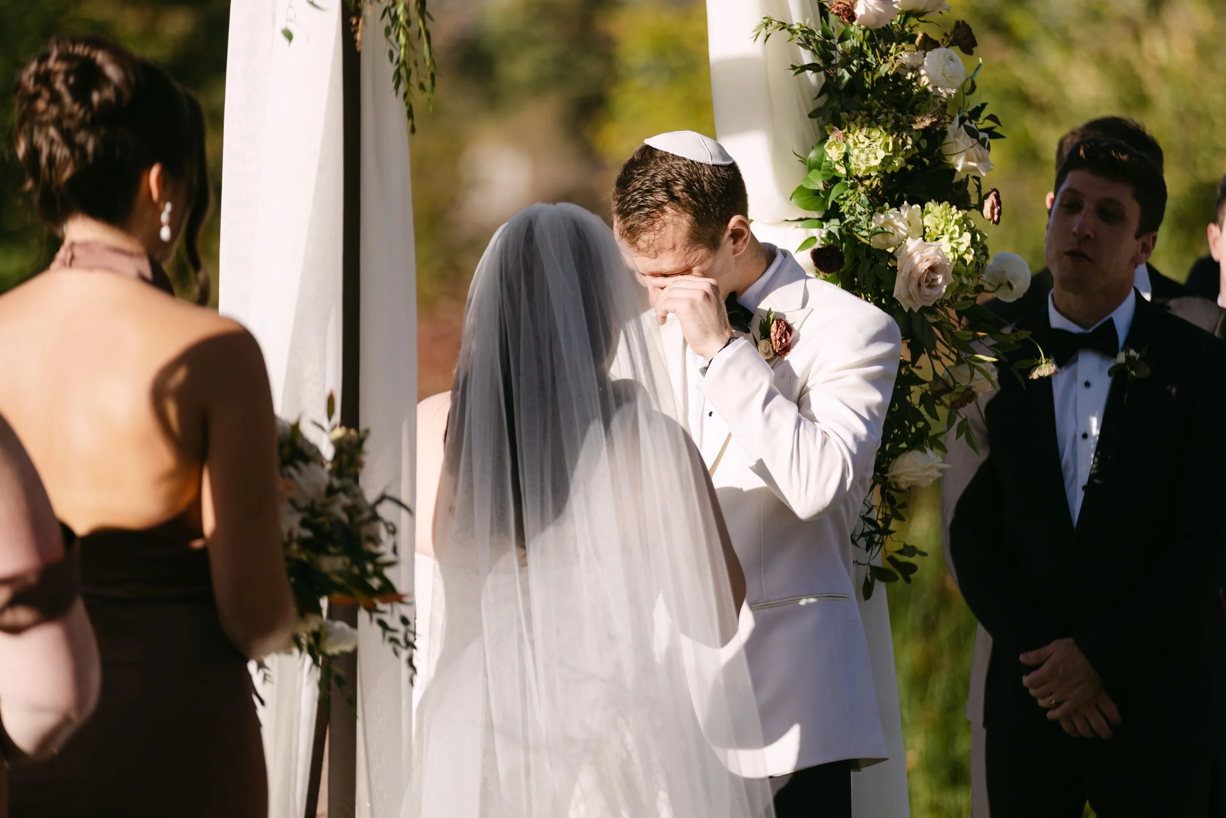 A groom in a white tuxedo and a Jewish kippah is emotional during his wedding ceremony, standing under an outdoor wedding arch decorated with white and green flowers, as a bride in a bridal veil watches.