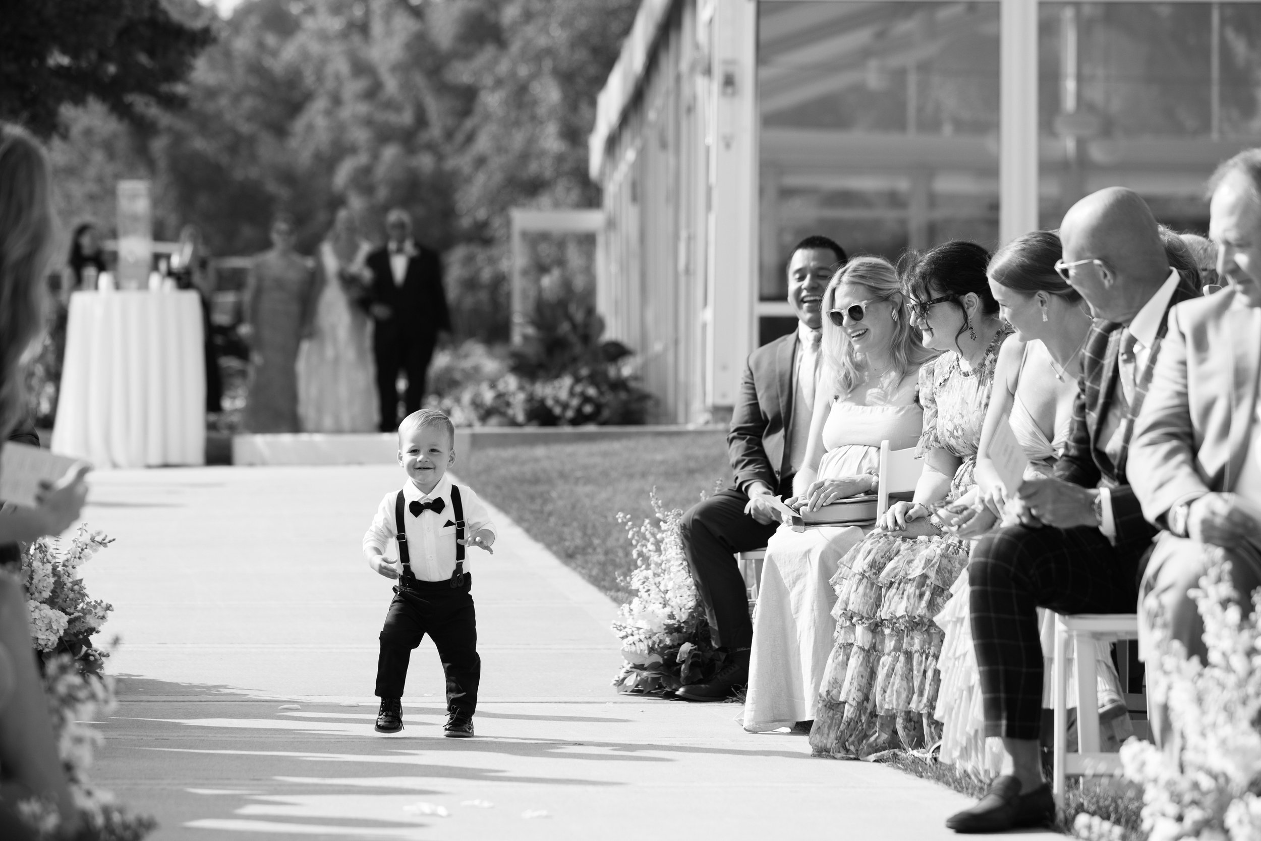 A young boy in a tuxedo running down a wedding aisle, smiling, with wedding guests seated on the right side watching and smiling in an outdoor wedding setting.