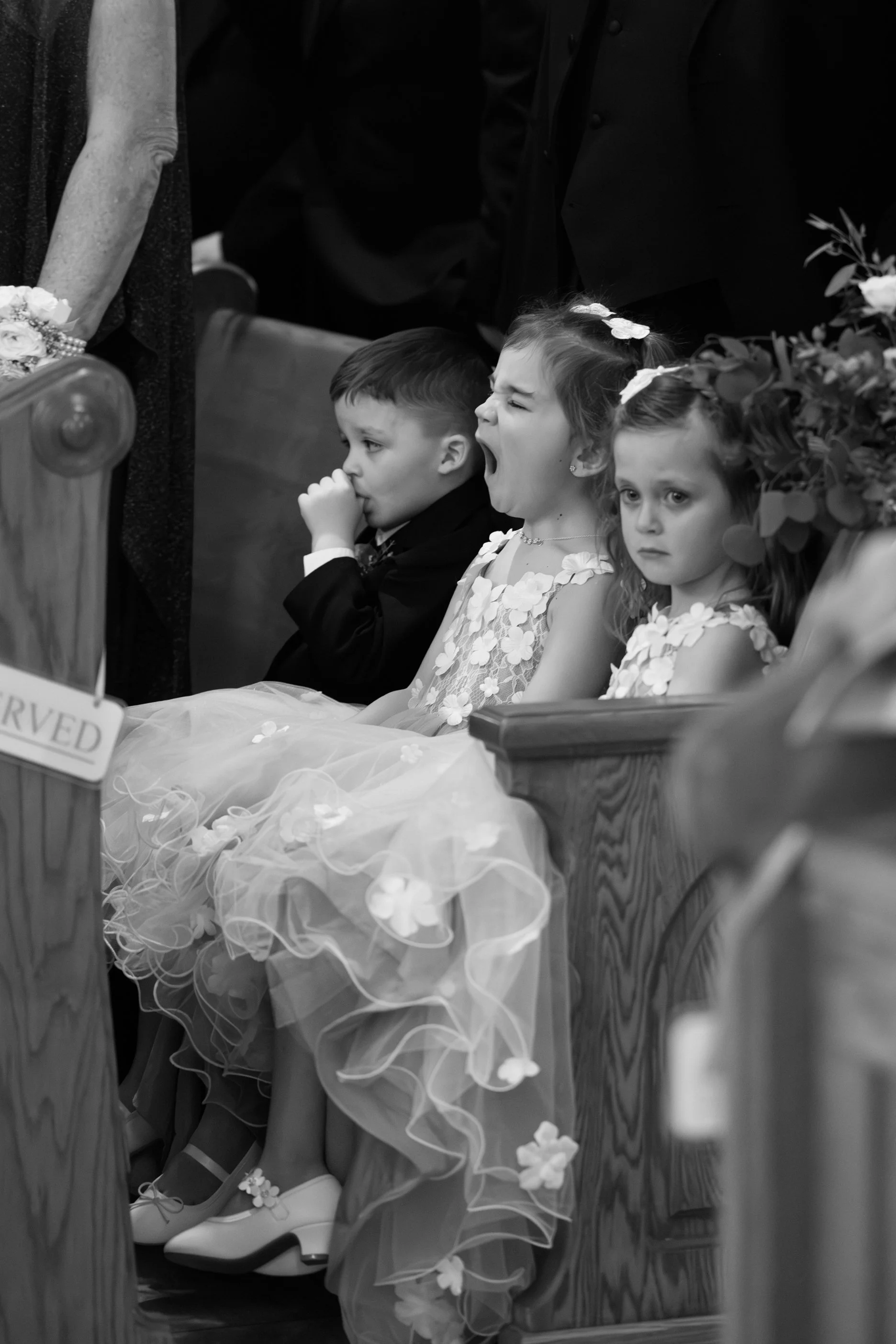 Three children, two girls and one boy, sitting in church pews during a ceremony; the girl in the center yawning, the other girl looking sad, and the boy looking thoughtful, all dressed in formal clothing.